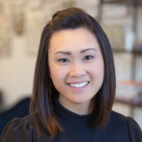 A woman with straight, shoulder-length dark hair, smiling, wearing a black top, indoors with a blurred background.