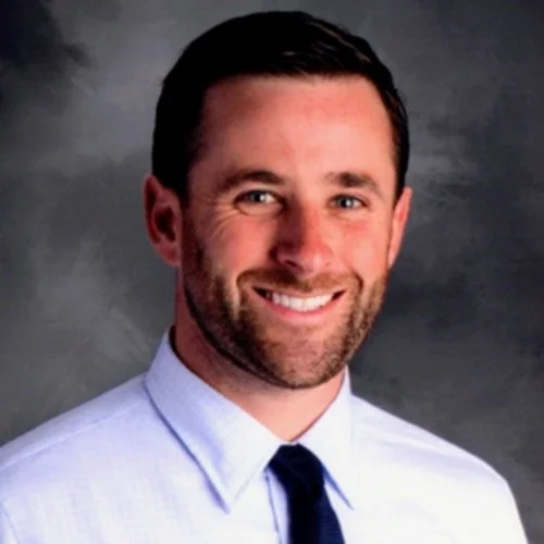 A smiling man with dark hair, wearing a light blue dress shirt and a dark tie, against a gray background.