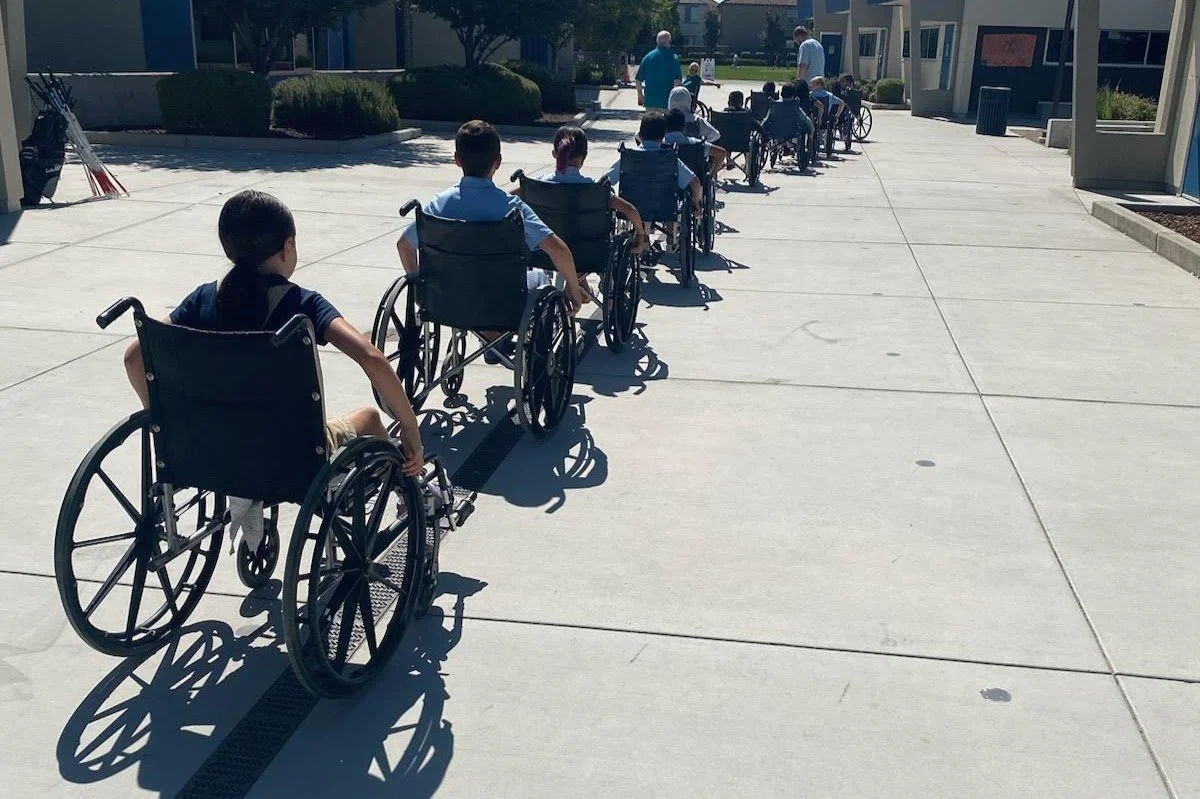 Line of children in wheelchairs outside on a sunny day, possibly at a school or care facility.