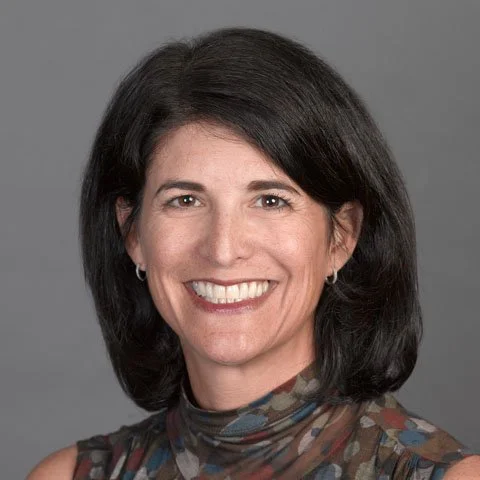 Portrait of a woman with shoulder-length dark hair smiling, wearing a patterned sleeveless top, against a gray background.