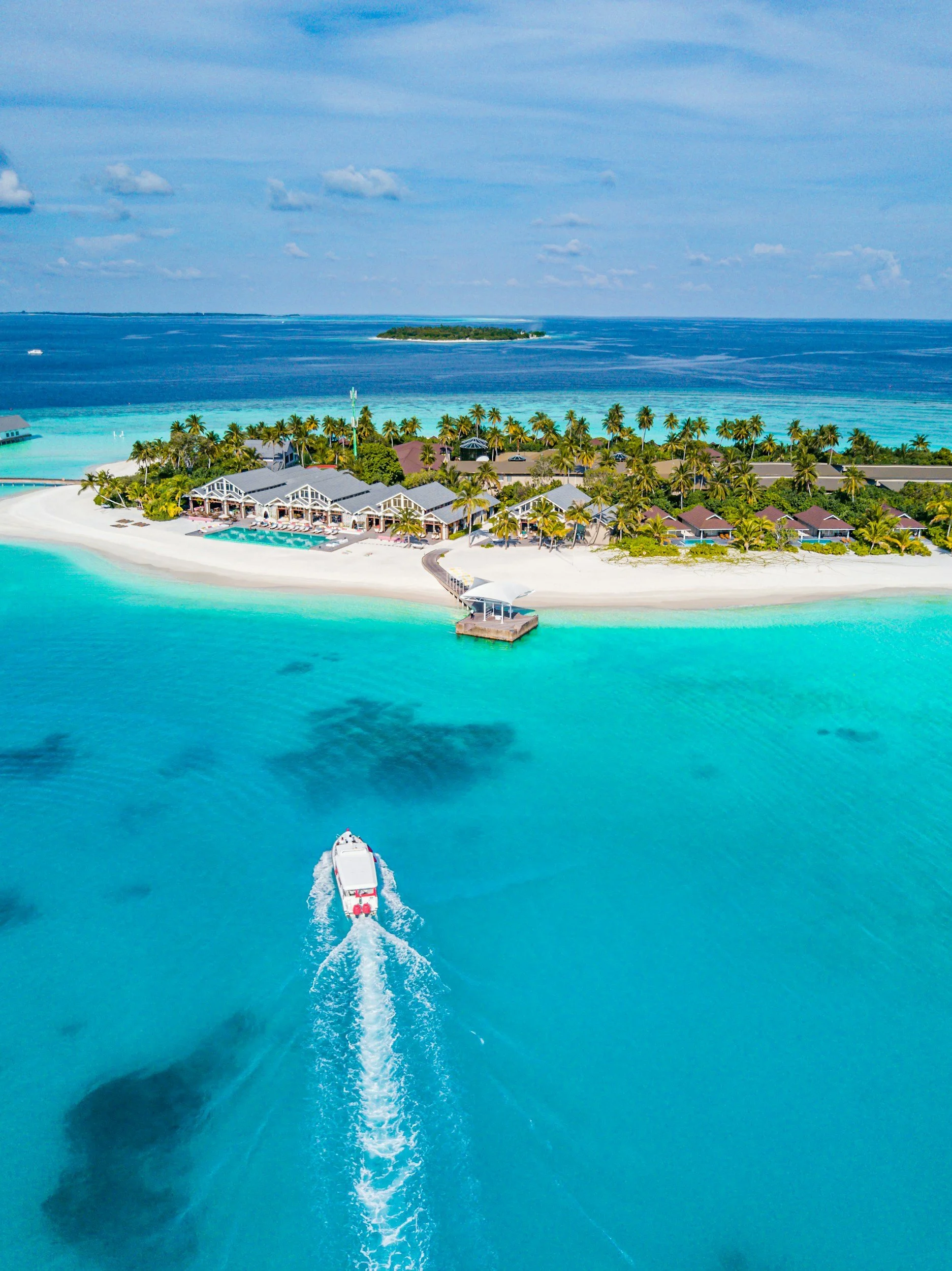 An aerial view of a luxurious tropical island resort with white sandy beaches, a swimming pool, palm trees, overwater bavilions, and a boat moving through clear turquoise water, with an ocean and a smaller island in the background.