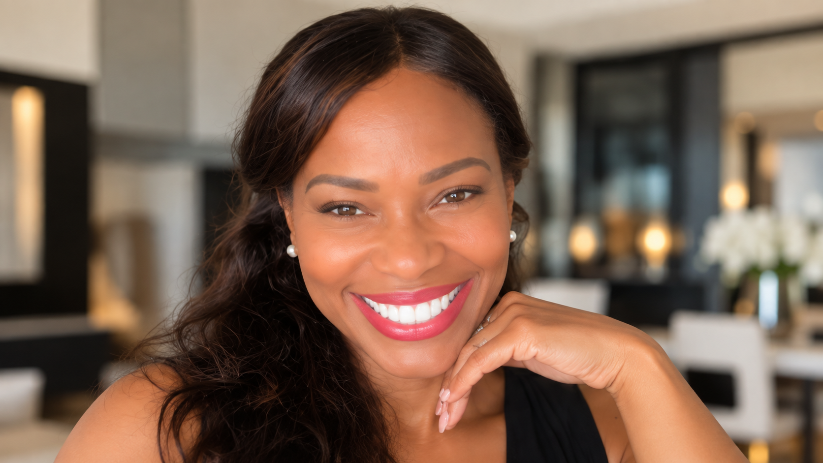 A woman smiling with bright white teeth, wearing pearl earrings, in an elegant modern interior.