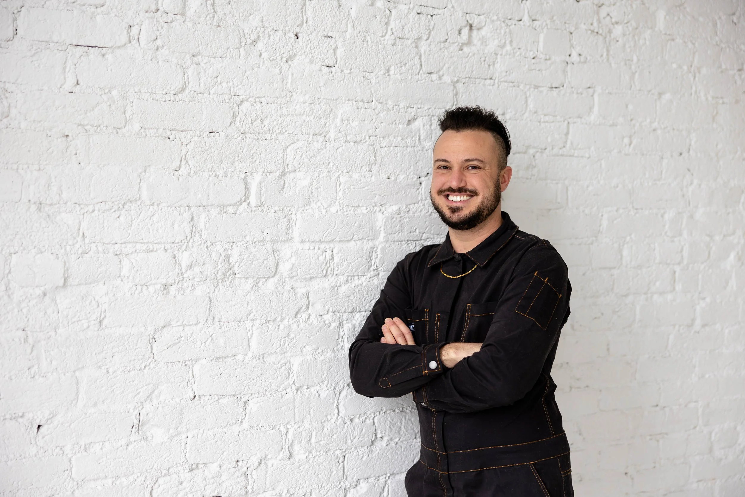 A man with a beard and dark hair smiling and crossing his arms, standing against a white brick wall.