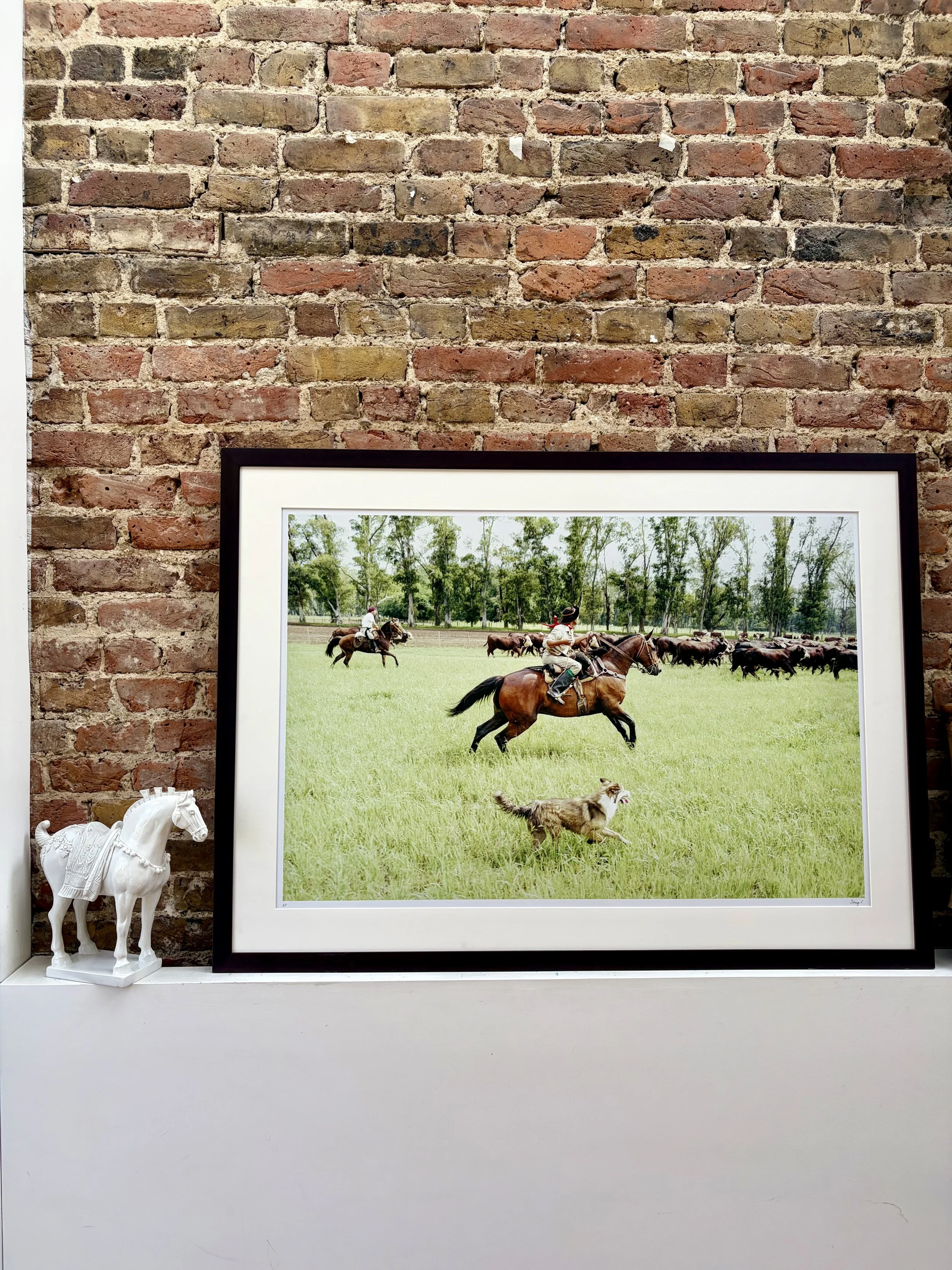 Photograph of a framed picture of a gaucho, his horse and his dog on a grassy field with trees in the background, positioned against a brick wall, with a small white horse figurine on a shelf below.