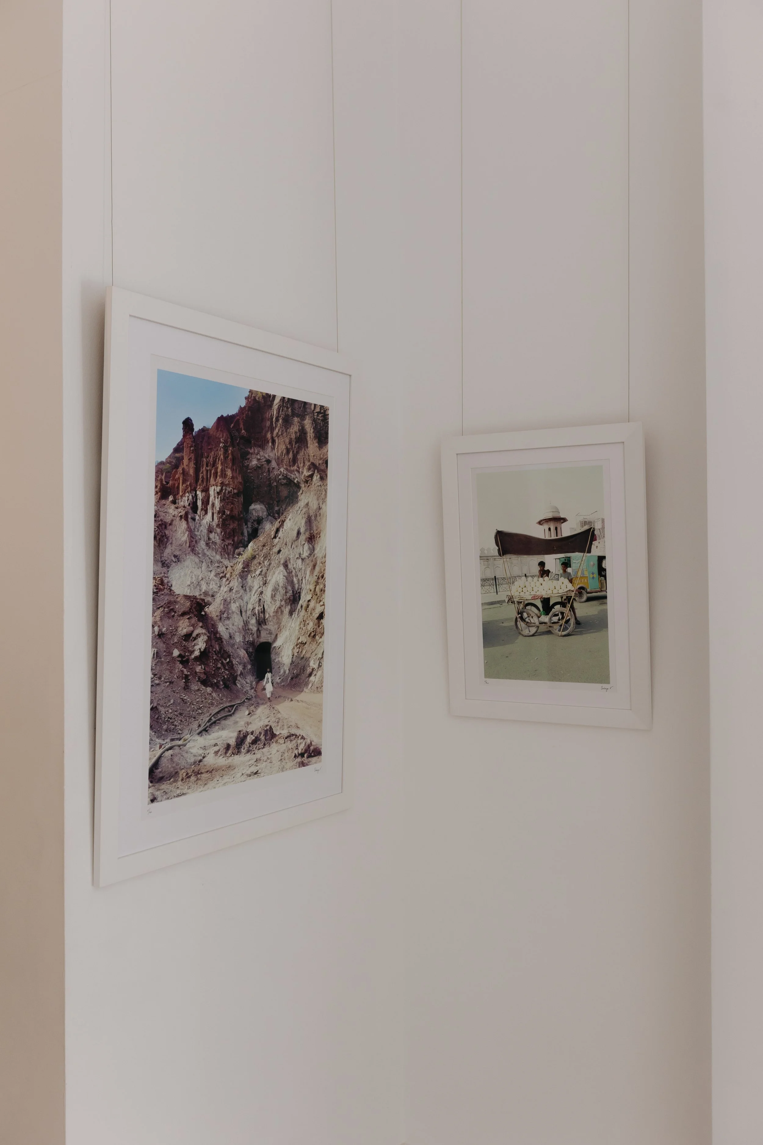 Two framed photographs hanging on a white wall, one depicting a mountain landscape and the other showing a street scene with a cart and a small building. bELONGING, sabaya verger first exhibition
