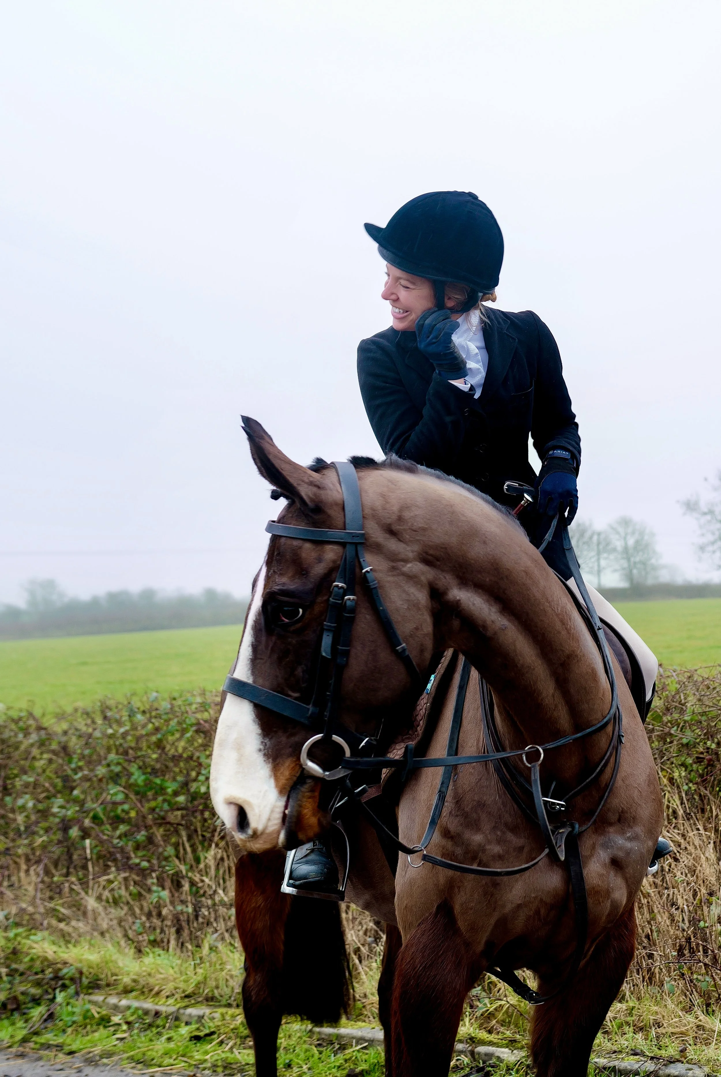 A woman, sabaya verger,  riding a brown and white horse outdoors on a cloudy day, wearing a black helmet, black jacket, gloves, and white shirt.