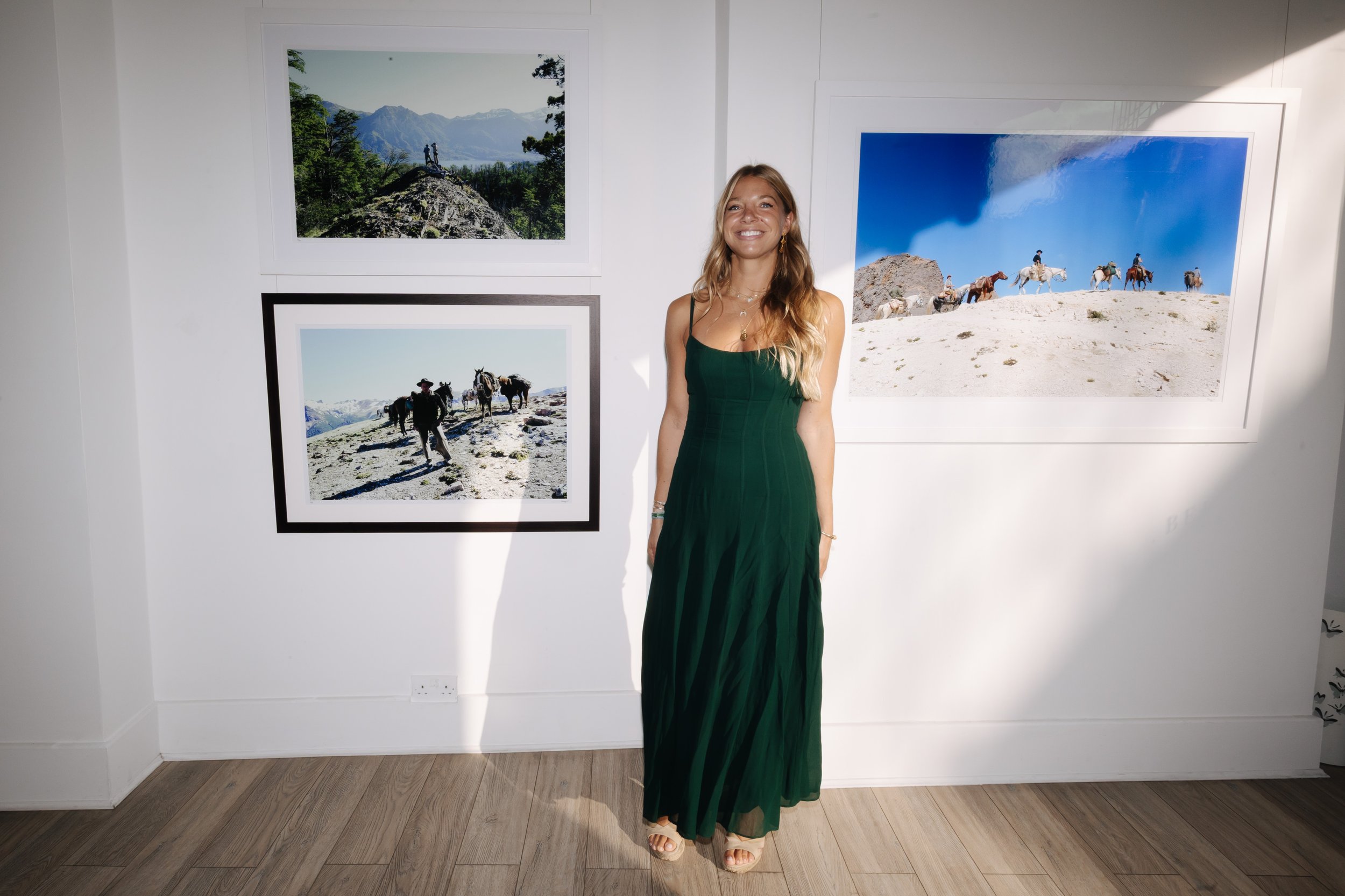 A woman in a green dress standing in an art gallery next to landscape photographs on a white wall. bELONGING, sabaya verger first exhibition