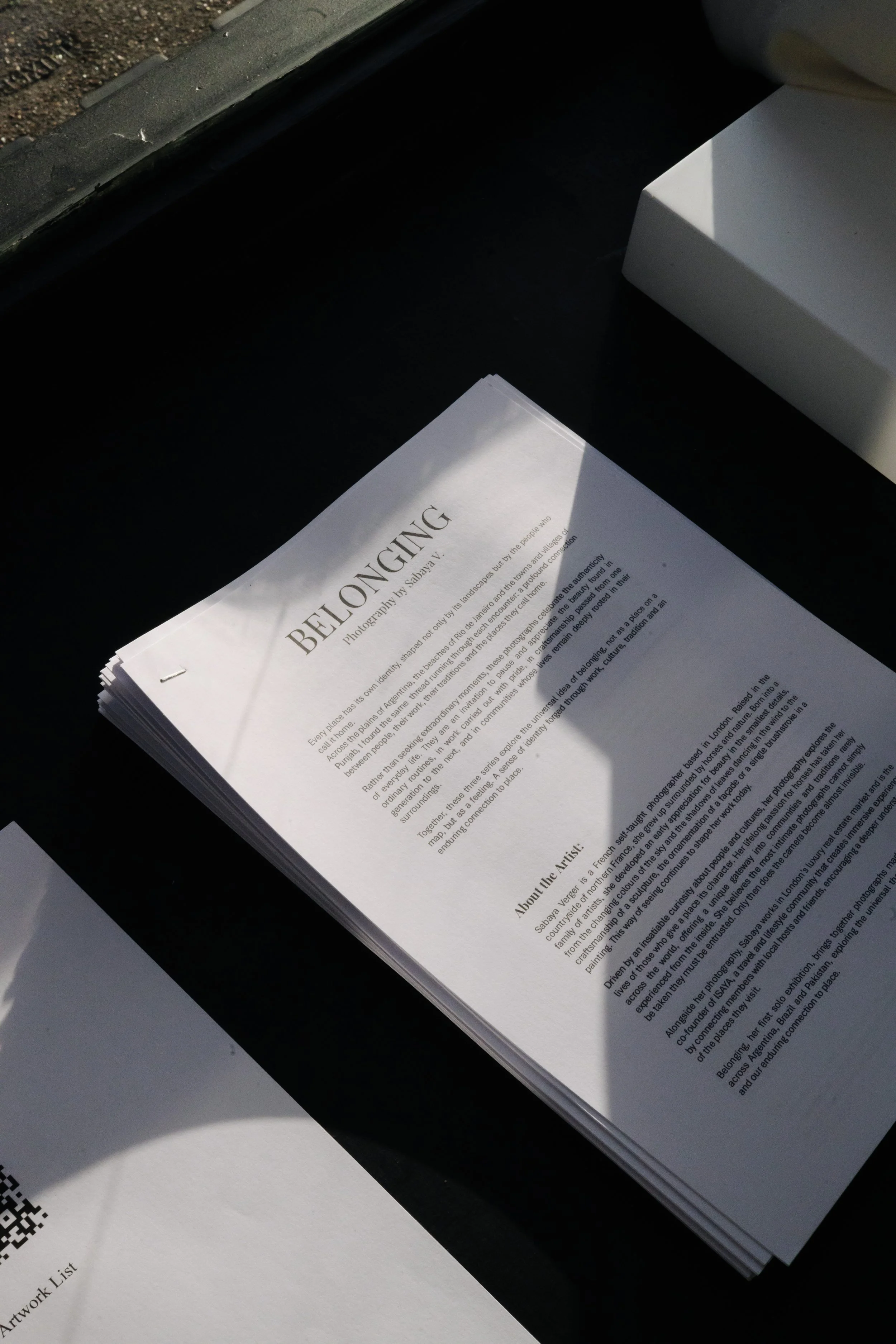 Close-up photo of printed pages containing text, titled 'BELONGING,' placed on a dark surface. Bright sunlight casting shadows on the pages and surface.