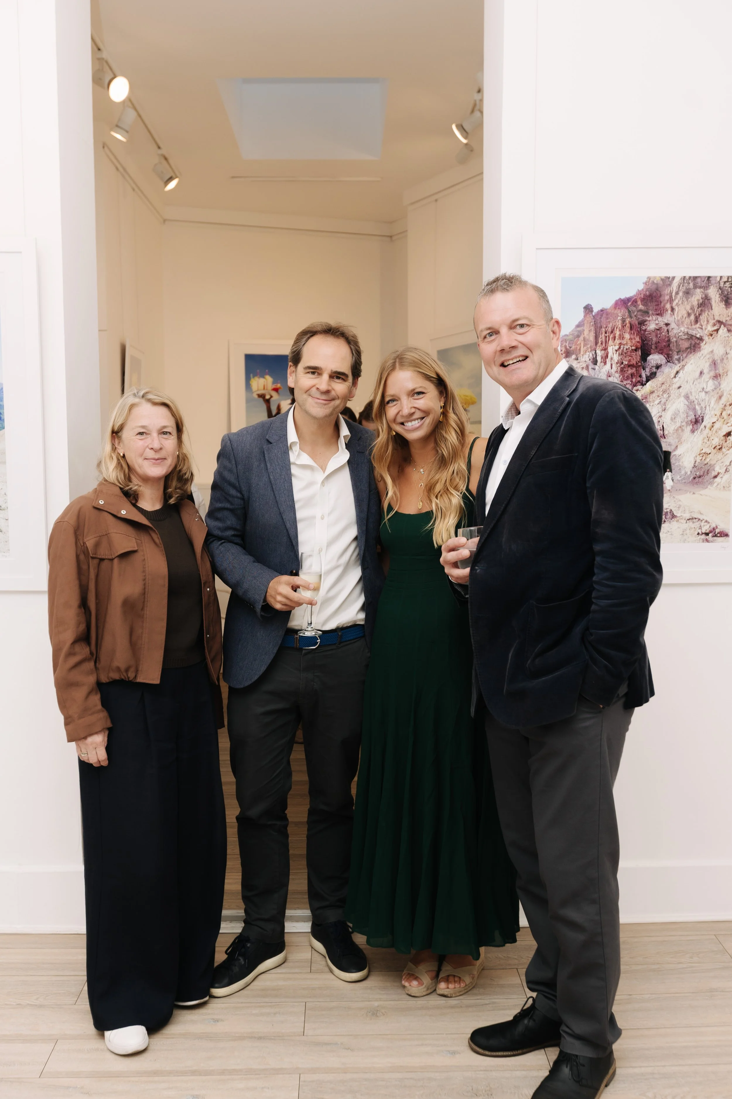 Four people standing in gallery with artworks on white walls, smiling at the camera. Two men in suits and two women, holding drinks, dressed semi-formally. sabaya verger and team Tedworth