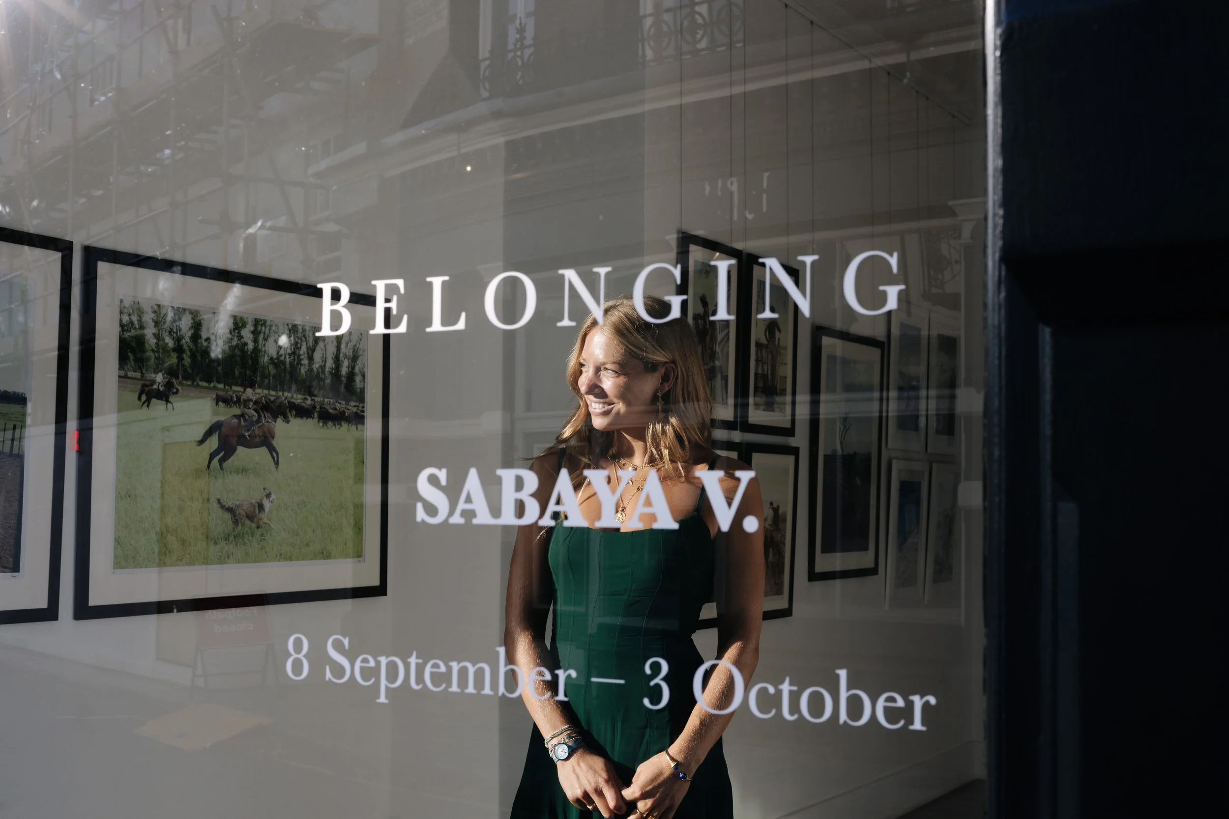 Sabaya Verg smiling  standing inside an art gallery with framed pictures of horses and countryside scenes on the wall behind her. The gallery window displays the text 'BELONGING' and event dates from September 8 to October 30. Her first exhibiton