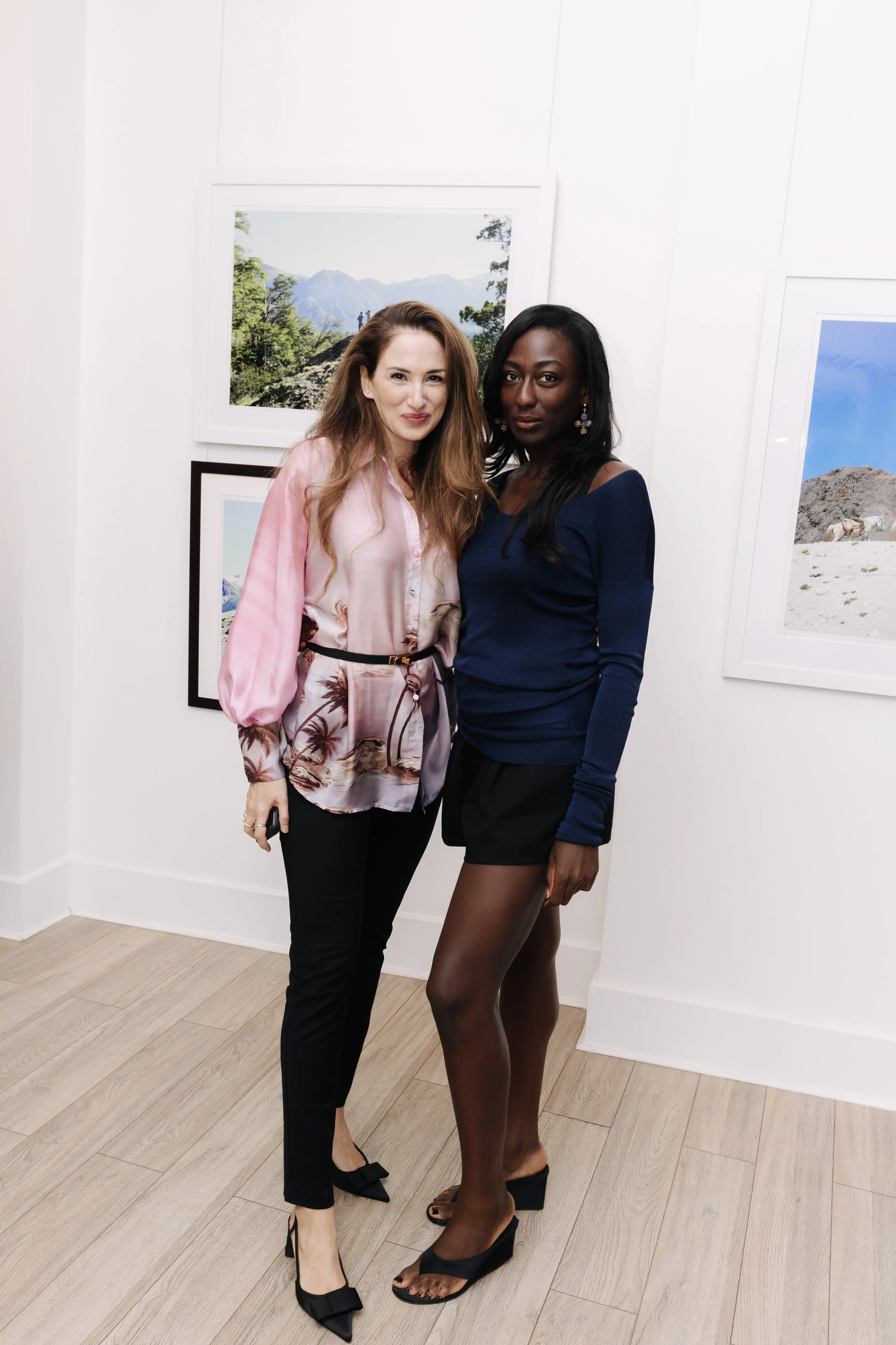 Two women standing close together, smiling, at an art gallery with framed landscape photographs on the white walls. One woman with light skin and brown hair is wearing a pink blouse and black pants, and the other with dark skin and black hair is wear