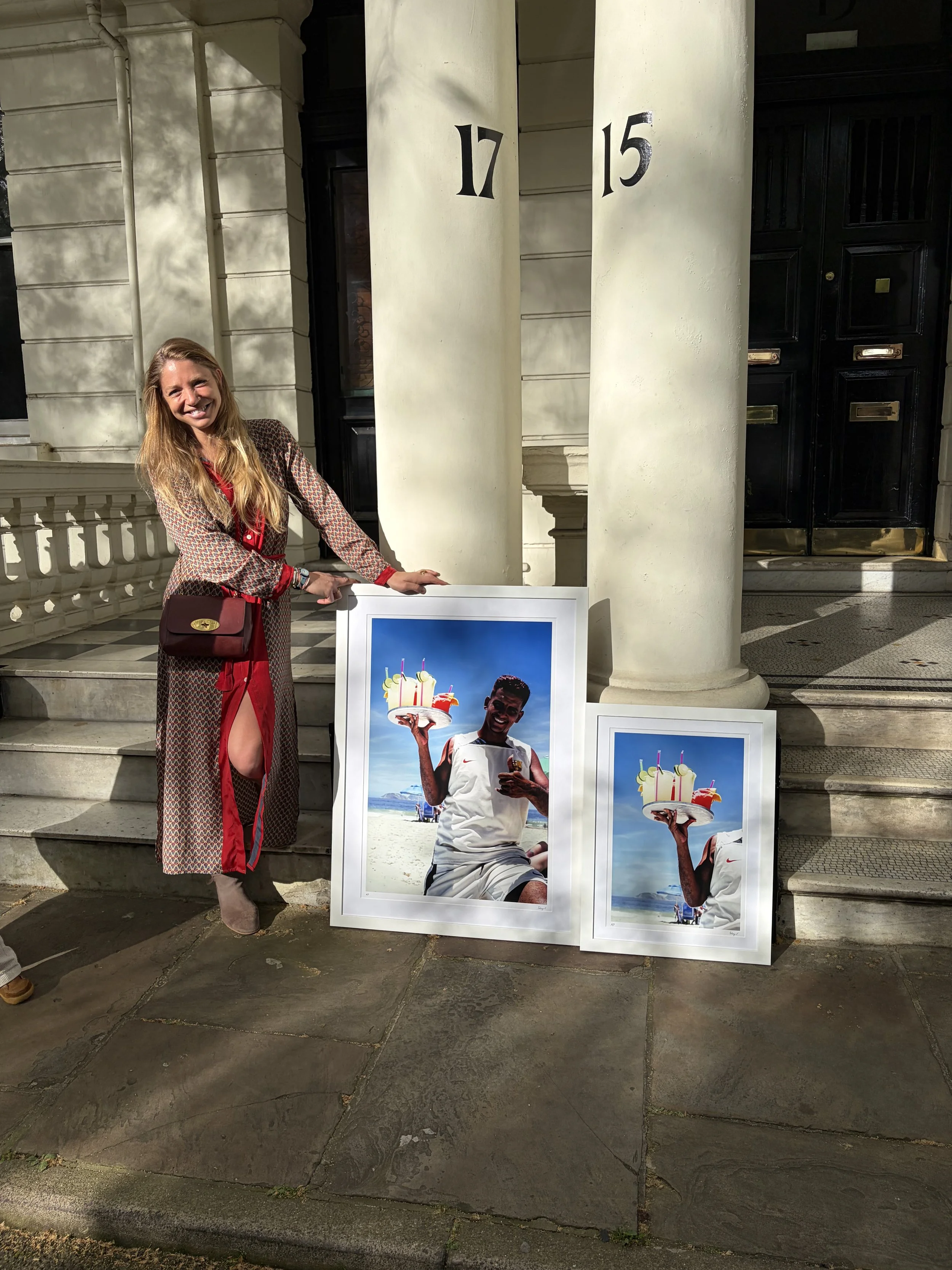 Sabaya Verger standing on stairs beside two large framed photographs; one of a man in white shorts holding a platter of coktails on Ipanema Beach in Rio de Janeiro, Brazil