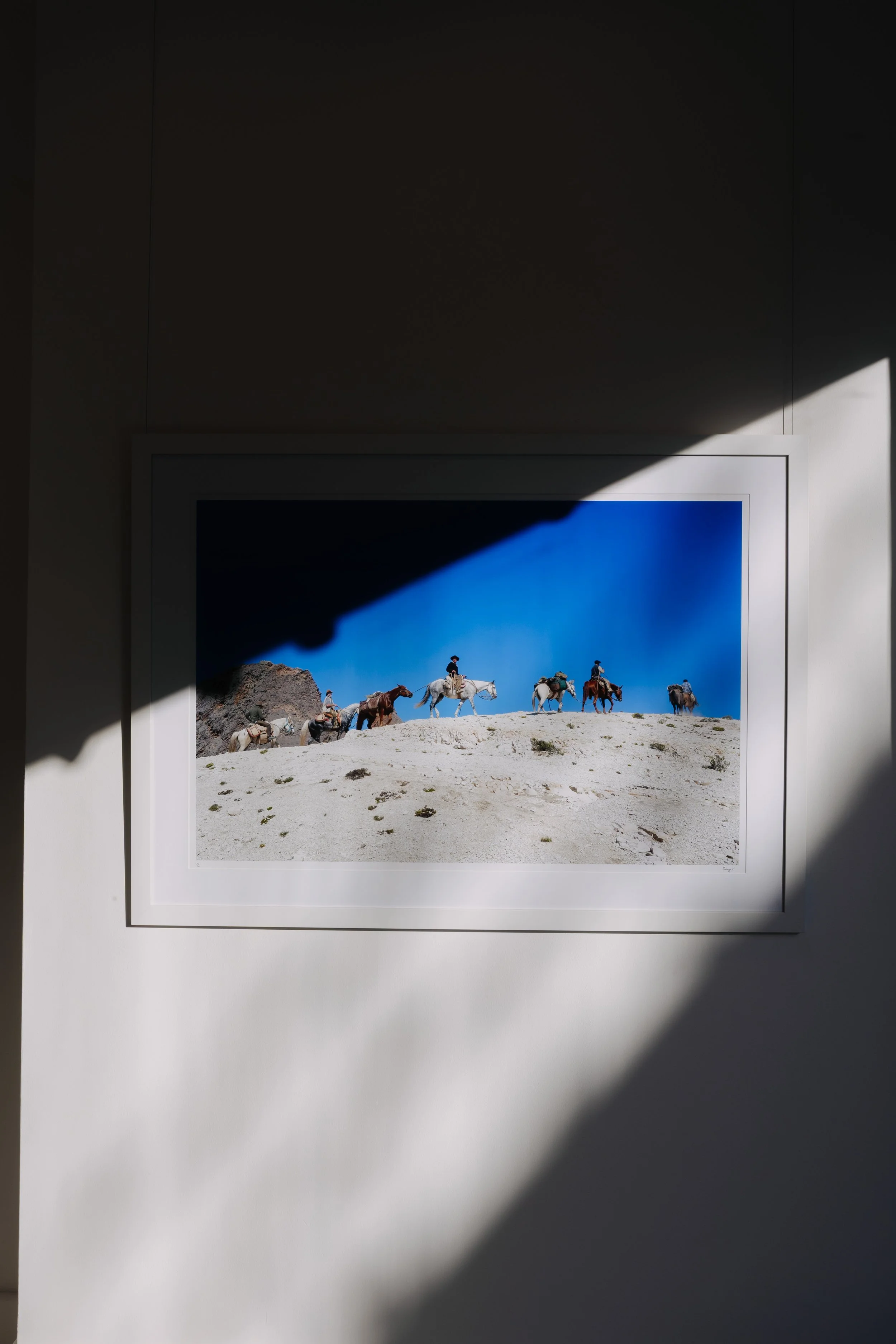 Photo of a framed picture of people riding horses in a desert landscape, sun casting a shadow over the top part of the picture, on a white wall. bELONGING, sabaya verger first exhibition Patagonia
