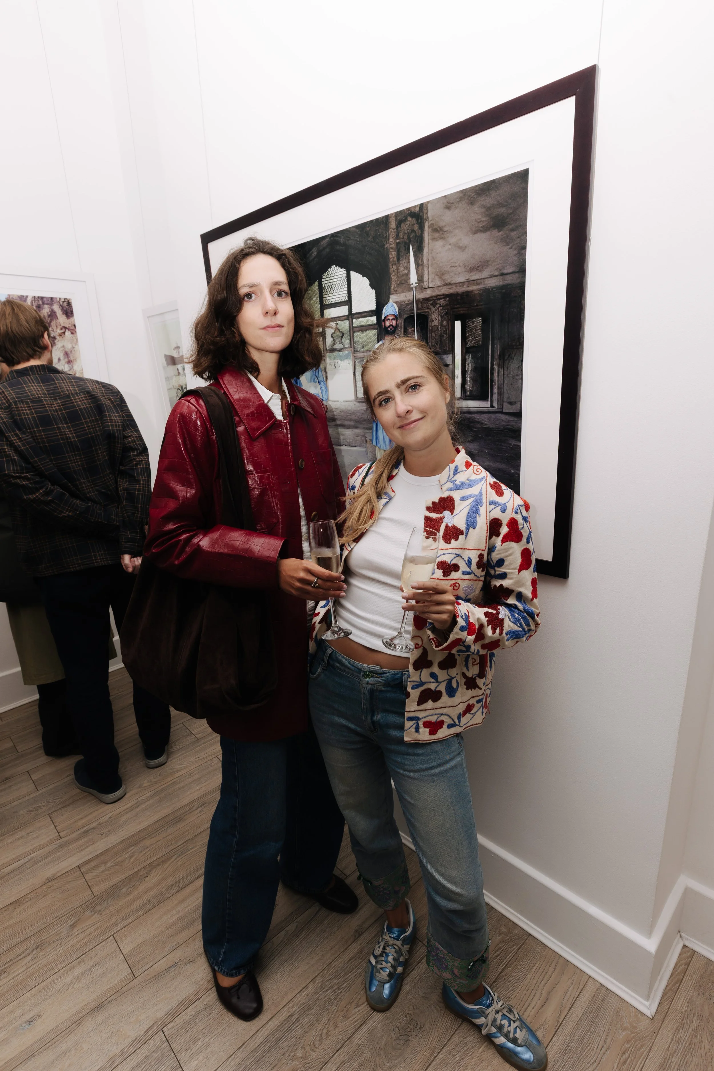 Two women standing close together in an art gallery, holding glasses of champagne, happiness and casual style, with a large framed photograph behind them. Simonetta Scheidt sabaya verger