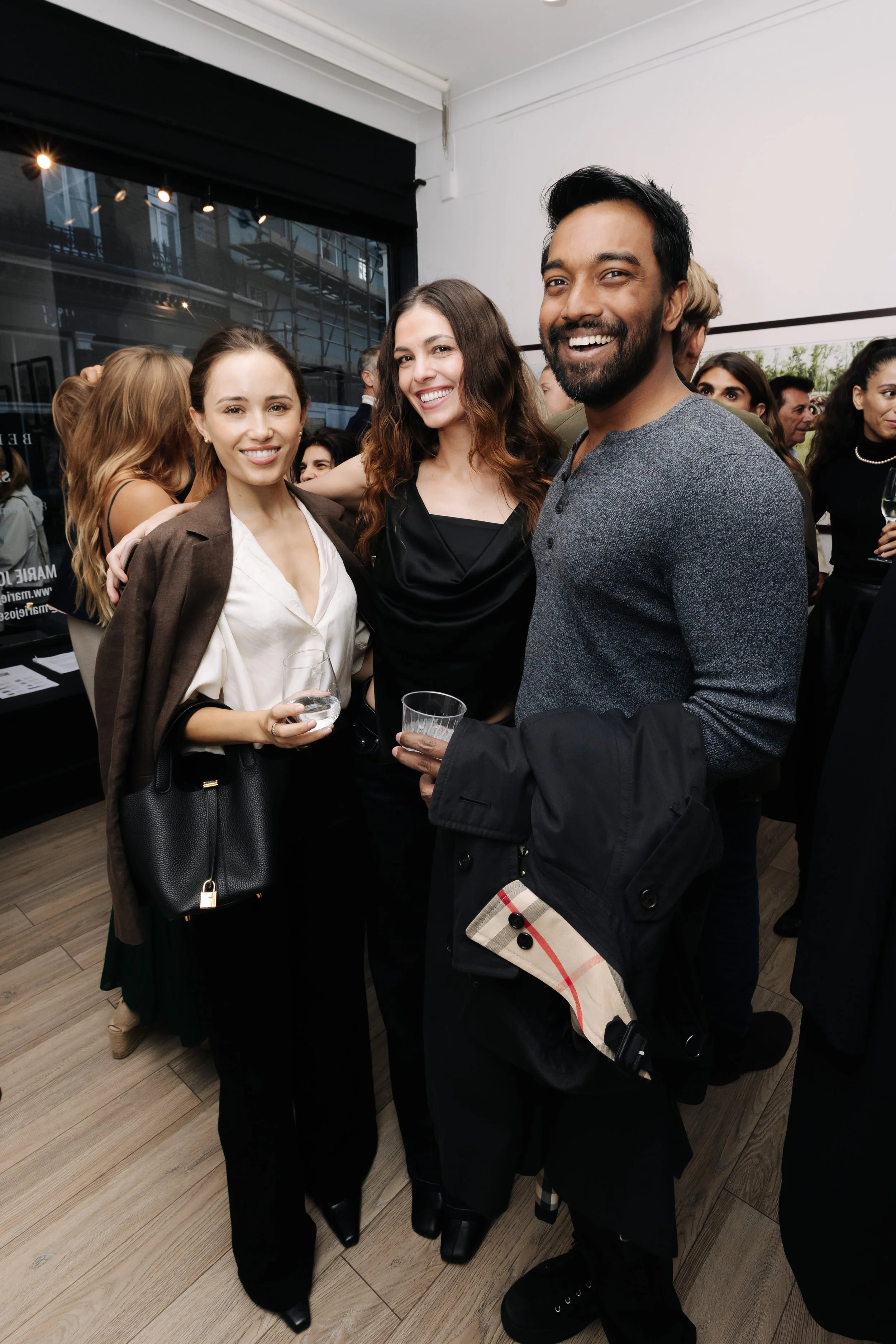 A group of people at a social event indoors, with three individuals in the foreground smiling and holding drinks, dressed in professional or semi-formal attire, with other guests mingling in the background. Kate Hutchins
