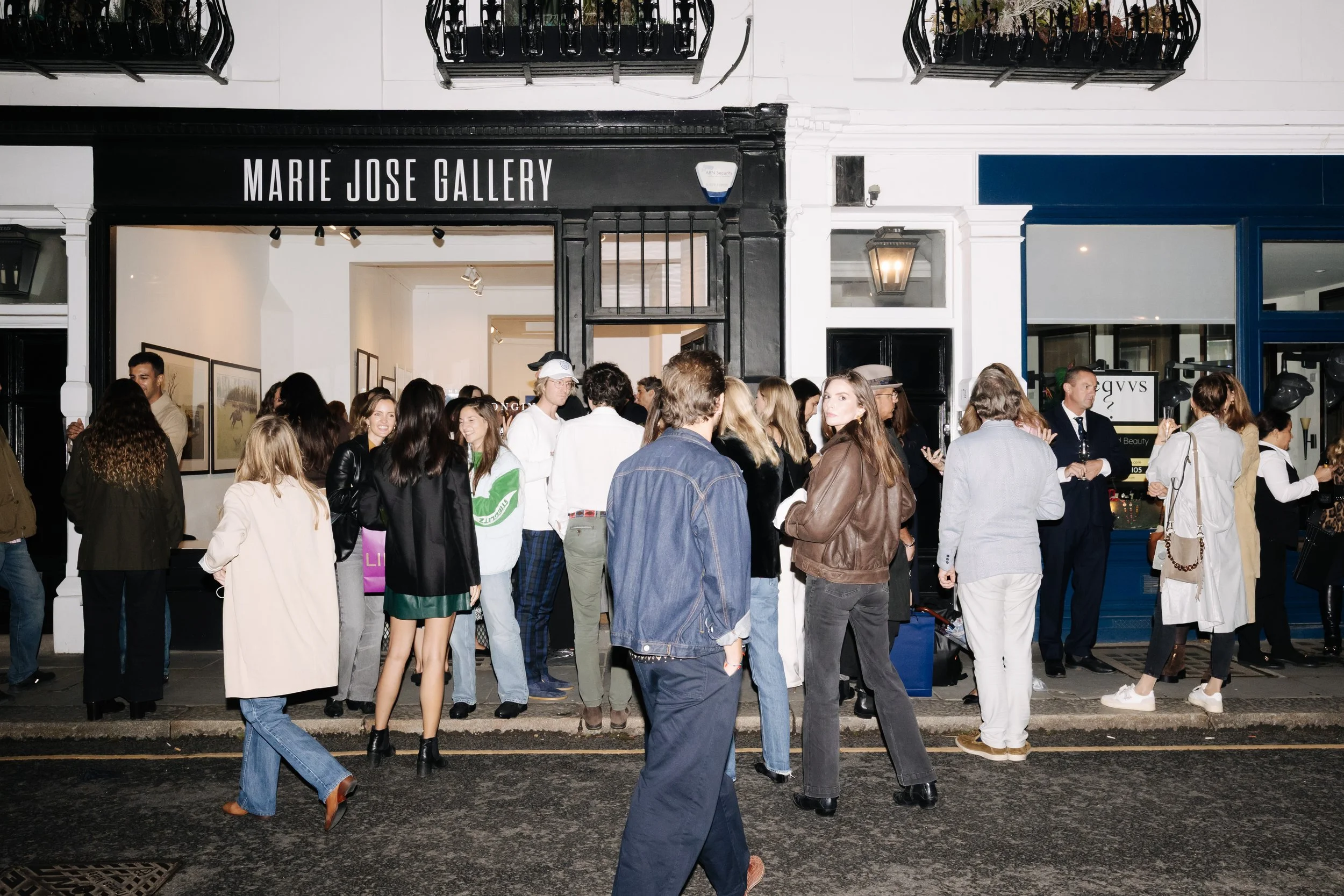 People gathering outside the Marie Jose Gallery, some engaged in conversation, others looking at their phones, with a blue storefront and framed artwork inside visible. bELONGING, sabaya verger first exhibition