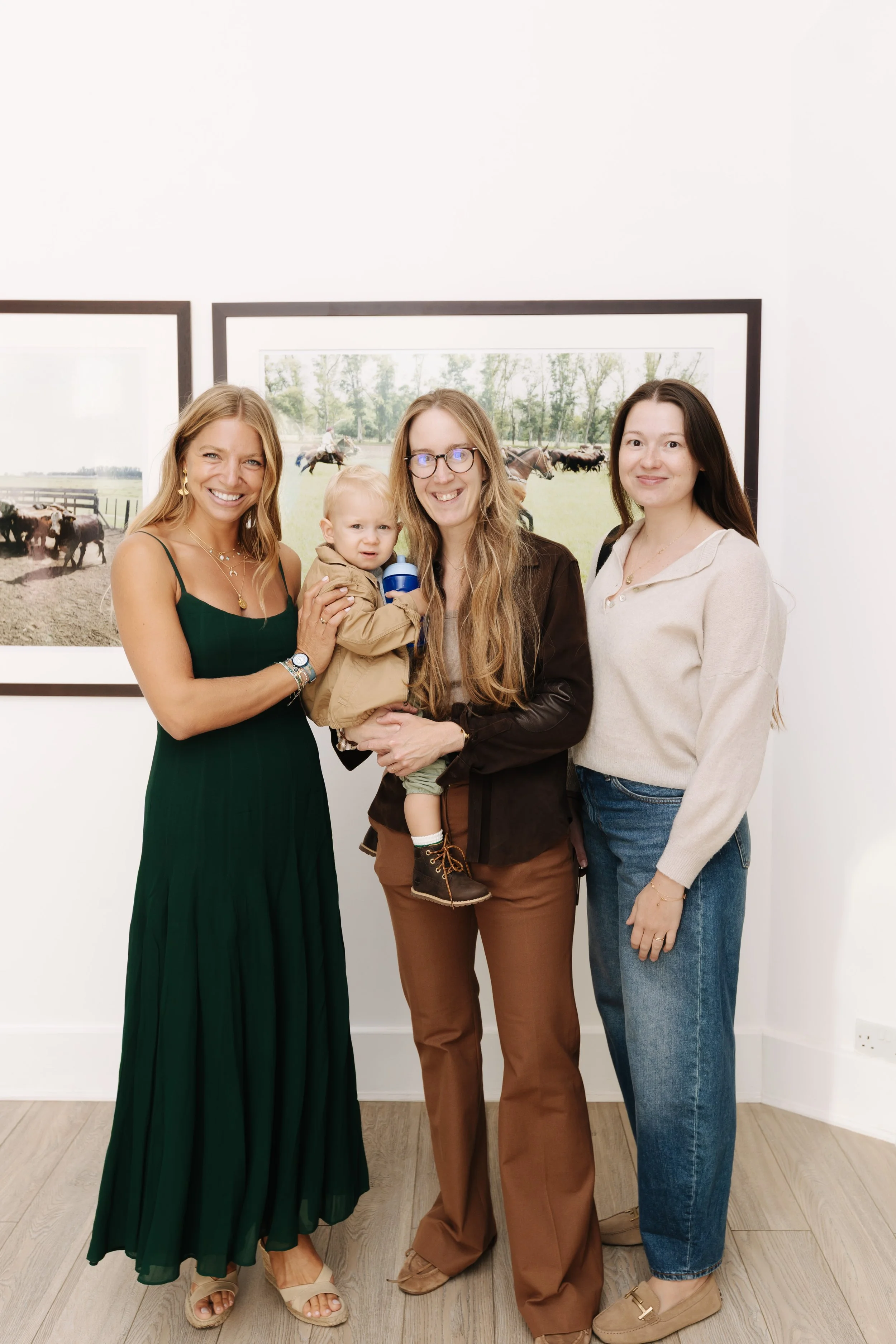 Four women and a young boy standing in front of wall art depicting cattle grazing in a field, smiling and posing for a photo. bELONGING, sabaya verger first exhibition Ines dawson