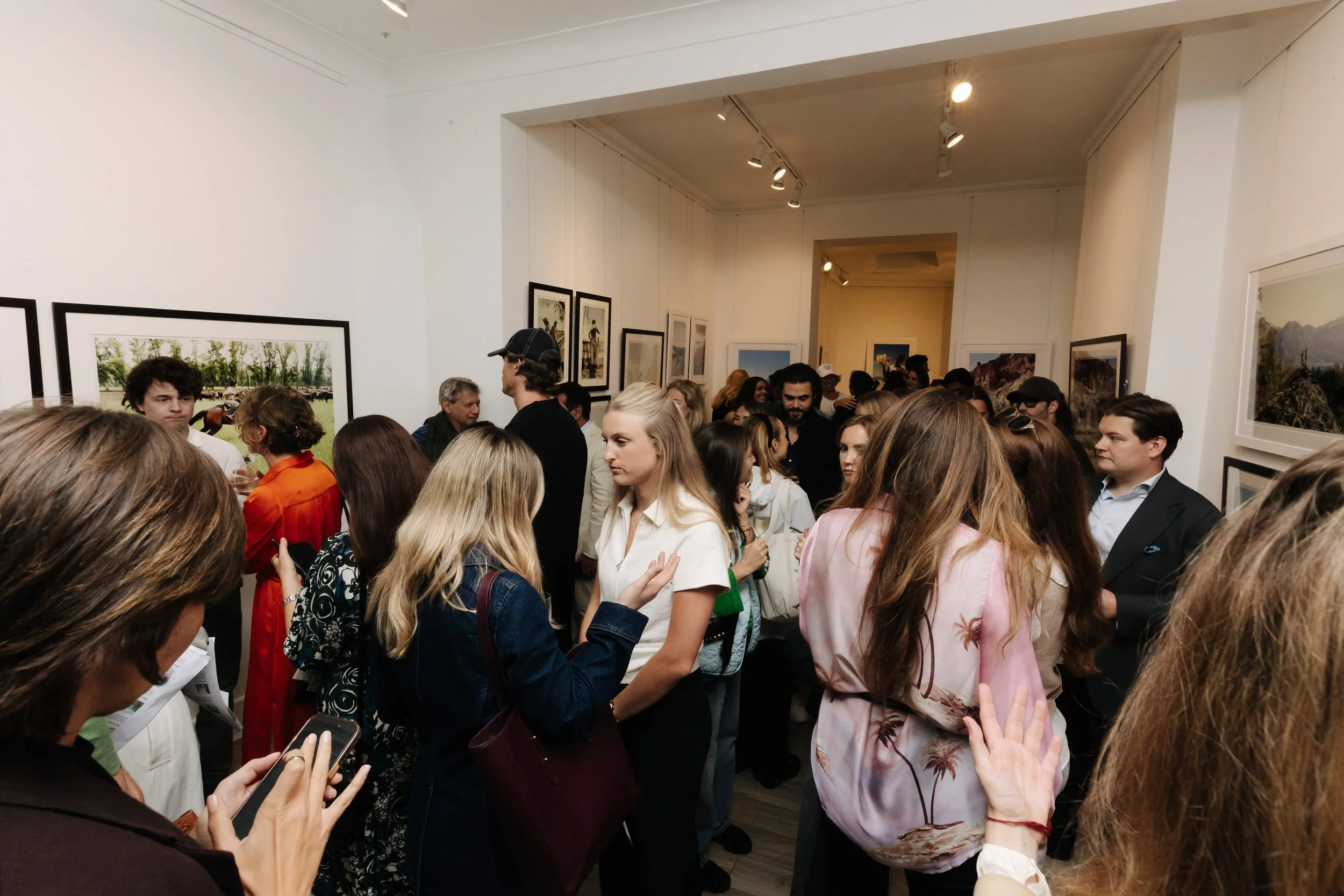 A crowded art gallery with people viewing paintings on white walls. The gallery has bright lighting and a diverse group of individuals engaged in conversations and looking at artwork. bELONGING, sabaya verger first exhibition