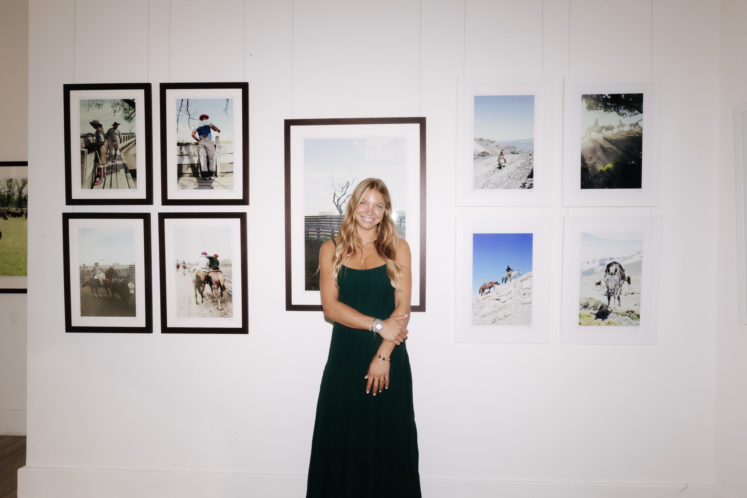 A young woman with long blonde hair wearing a dark green dress, smiling and standing in front of a white wall with framed landscape photographs of mountains, horses, and rural scenes. bELONGING, sabaya verger first exhibition