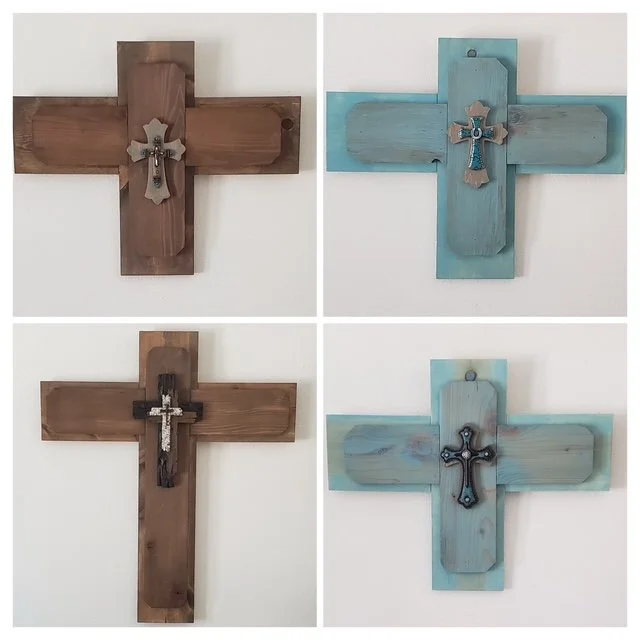 Four wooden crosses mounted on a white wall, each with a different design and color scheme, including natural brown and painted light blue, featuring various decorative crosses attached.