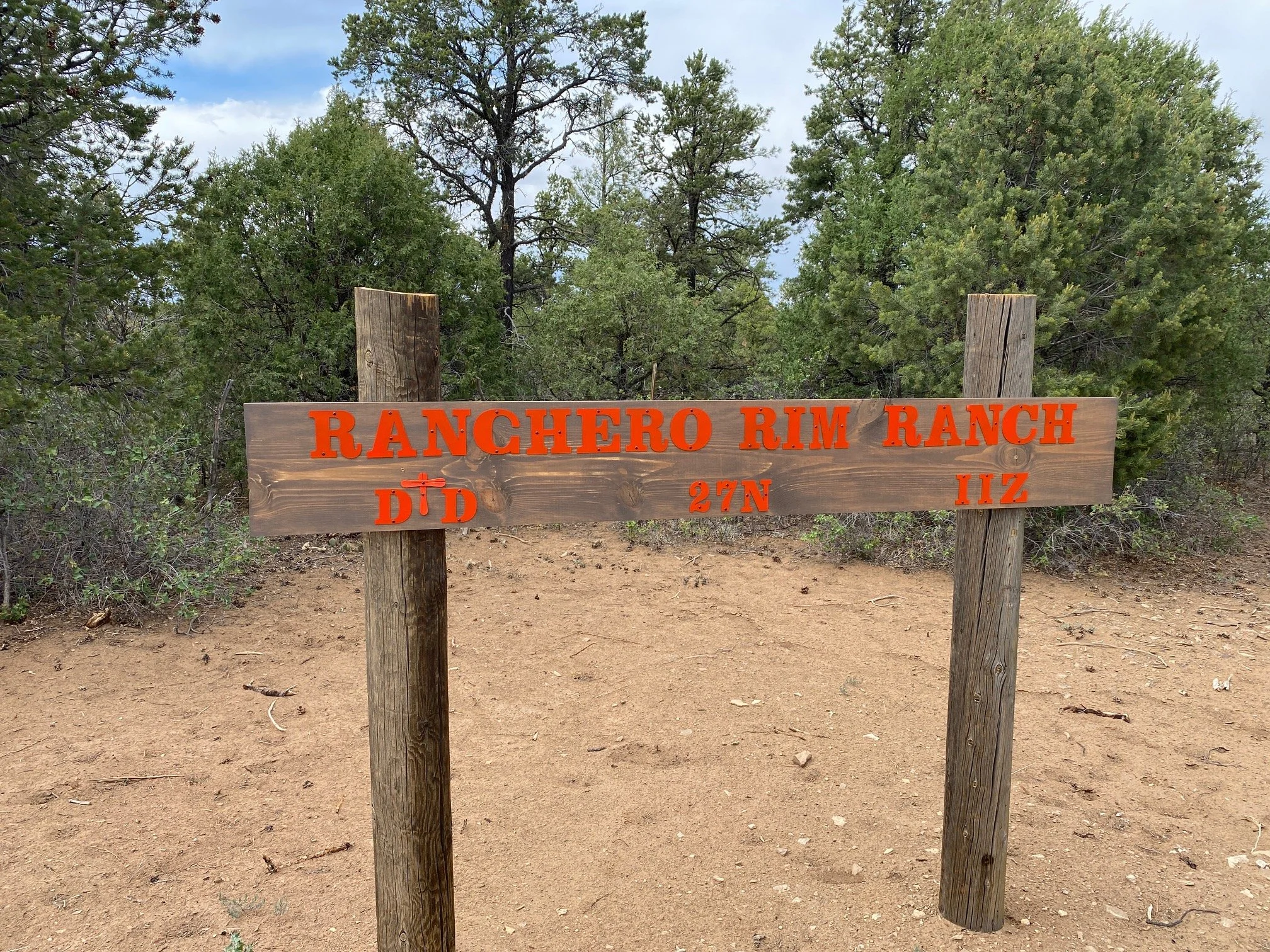 Wooden sign reading 'Ranchero Rim Ranch' with directional information, situated on dirt ground surrounded by dense green trees under a partly cloudy sky.