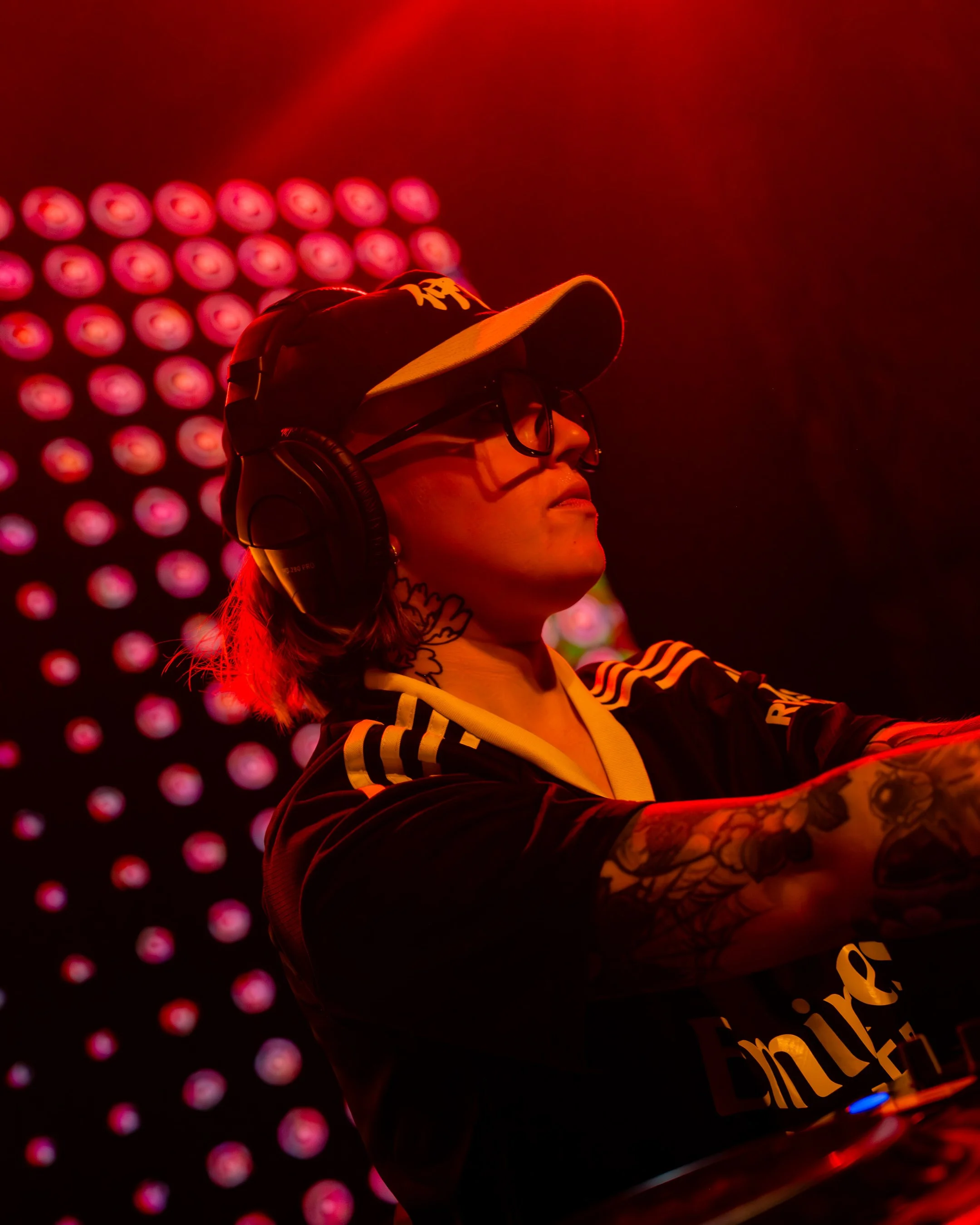 A woman wearing glasses, a baseball cap, and headphones, with tattoos on her arm, in a red-lit environment with a background of red circular lights.