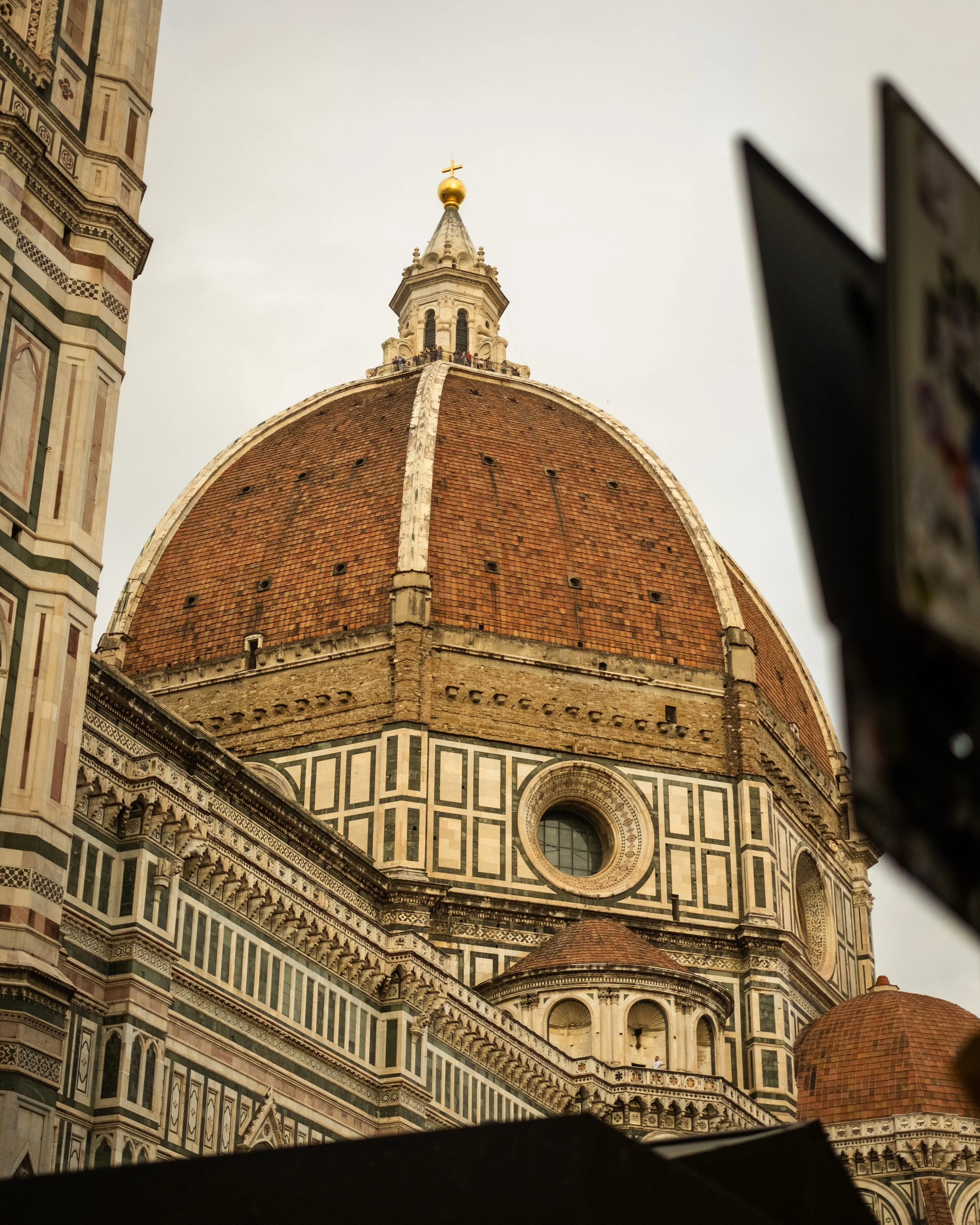 The image depicts the top part of an ornate cathedral with a large dome covered in reddish-orange tiles, topped with a smaller spire and a golden orb with a cross. The cathedral features intricate decorative patterns and columns, with a section of th