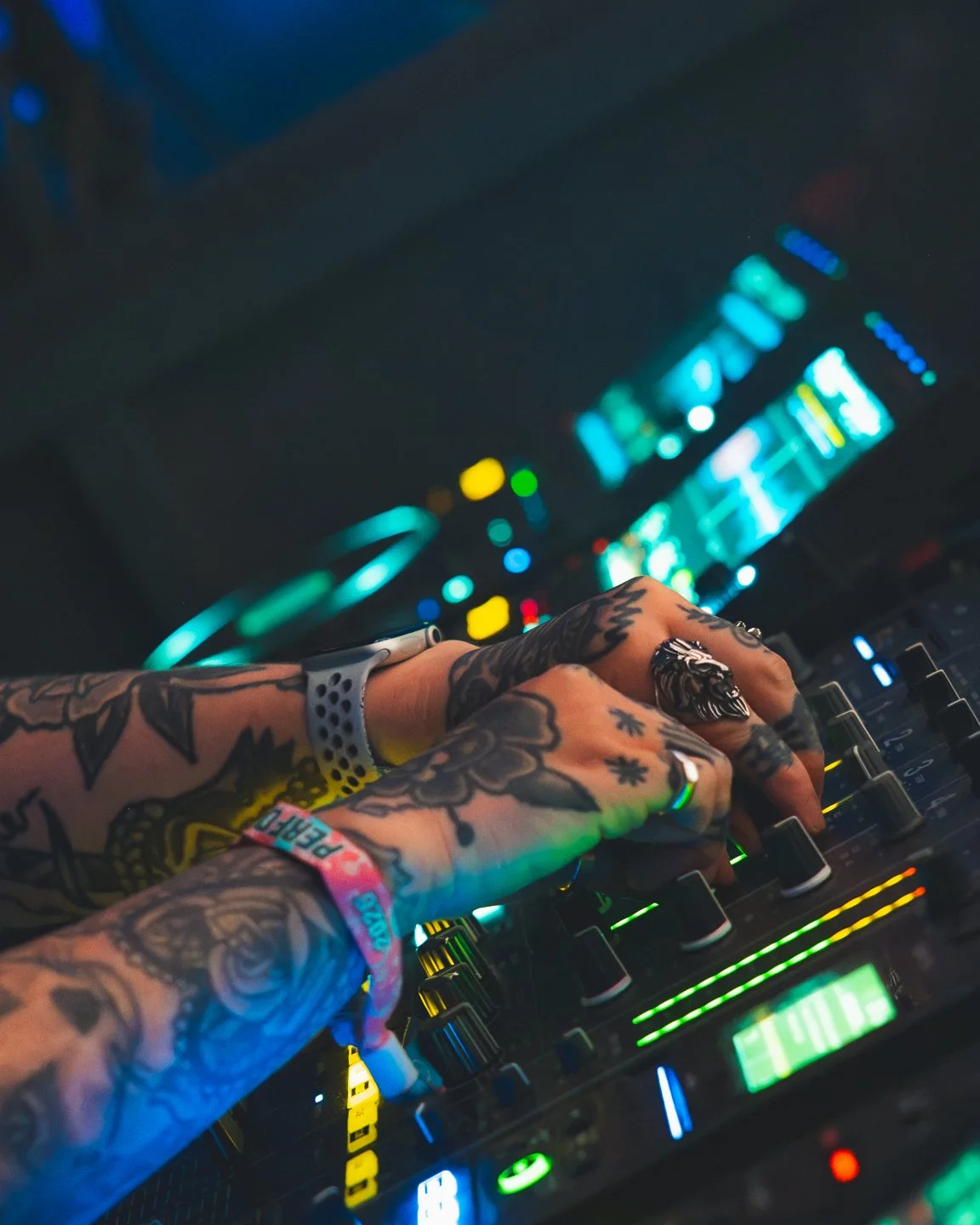 Tattooed hands operating a DJ mixer in a nightclub with colorful LED lights and screens in the background.