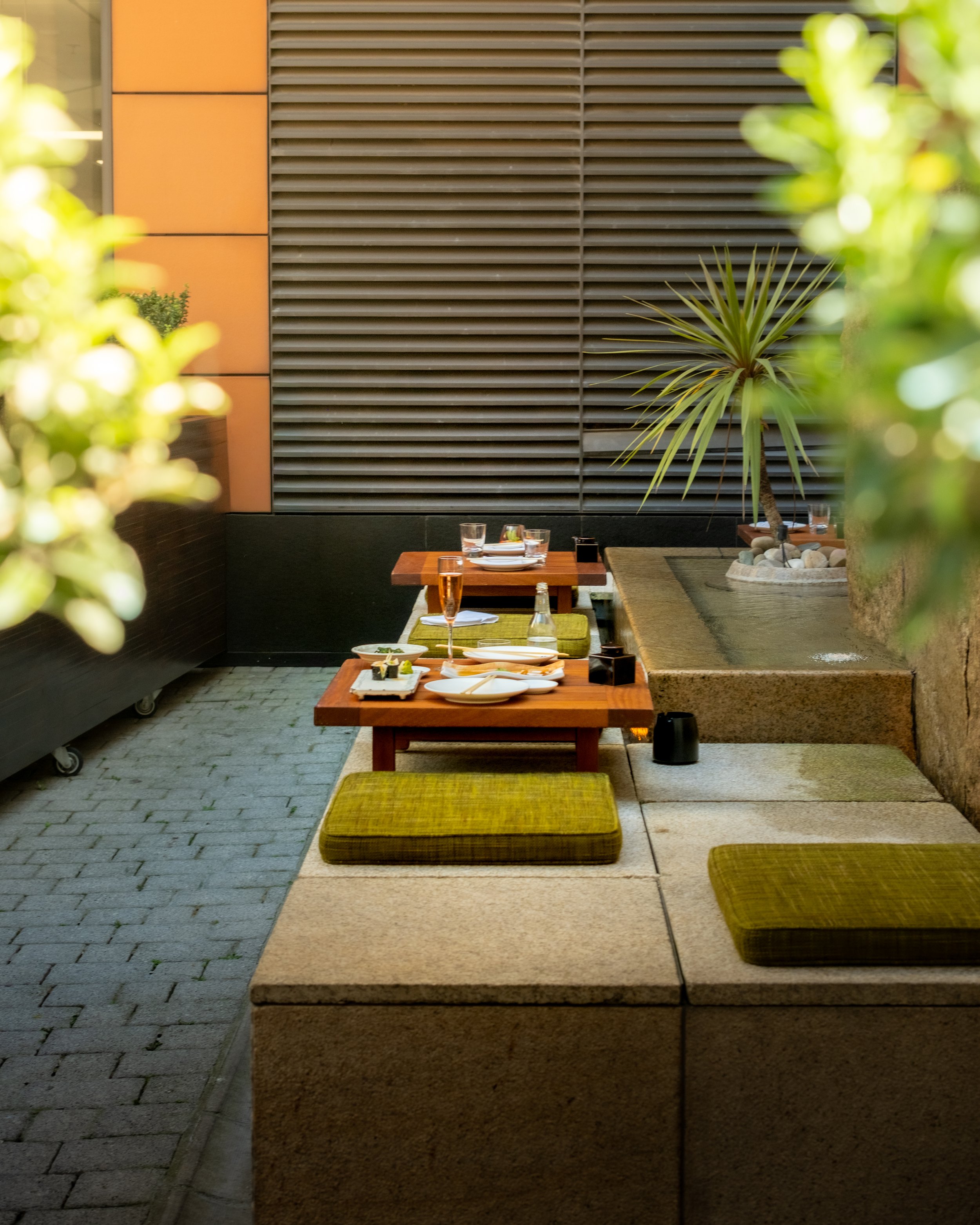 Outdoor dining area with low tables, yellow cushions, and surrounding greenery, including a small palm tree and plants, in front of a building with gray slatted walls and orange panels.