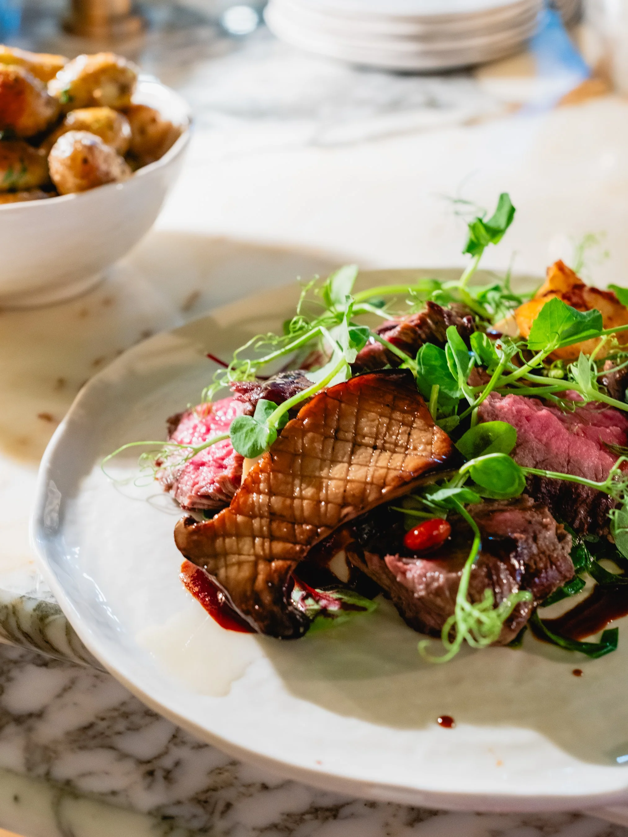 Close-up of a plated dish featuring sliced cooked beef topped with grilled scallops, garnished with microgreens, on a white plate with a marble countertop in the background.