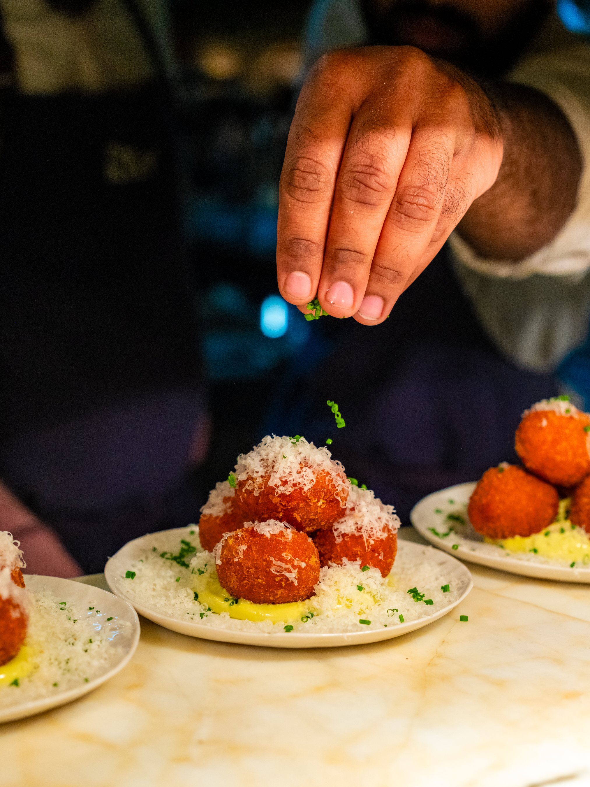 A person sprinkling chopped green herbs onto a plate of fried food ball appetizers with shredded cheese and a sauce base.