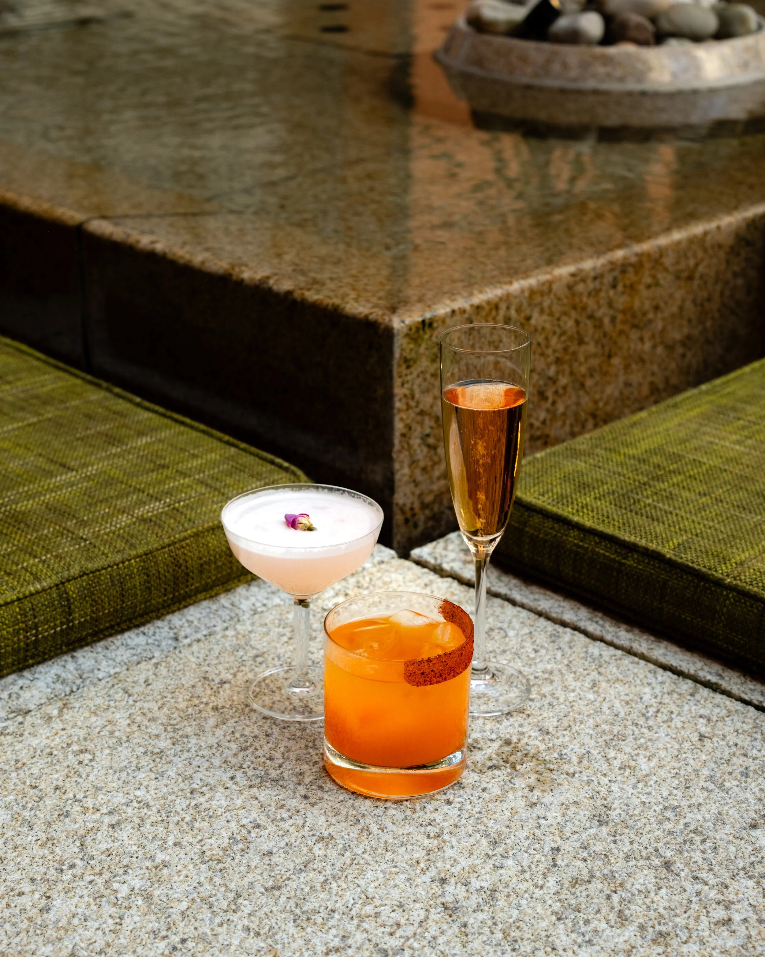 A close-up of a granite countertop with three glasses of cocktails, one orange, one pink, and one with a dark liquid, with a potted rock garden in the background.