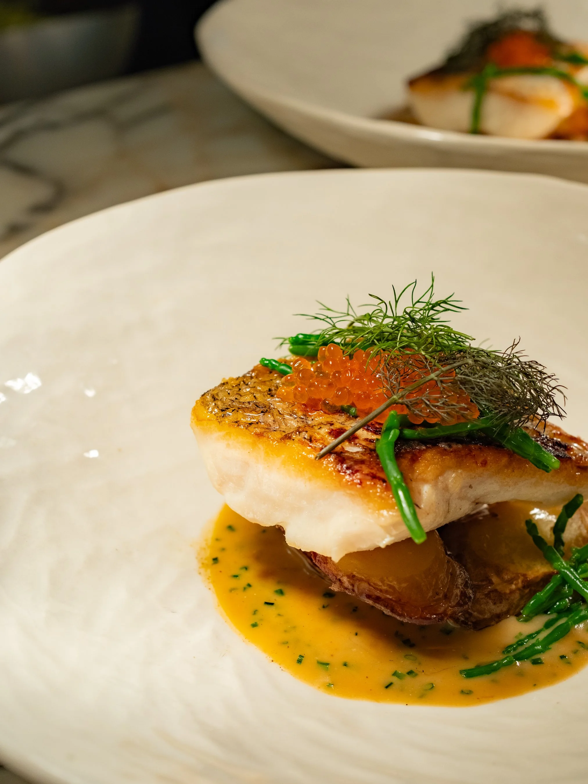 A plated gourmet dish featuring a seared fish fillet topped with orange fish eggs, fresh herbs, and green vegetables, served with a light sauce.