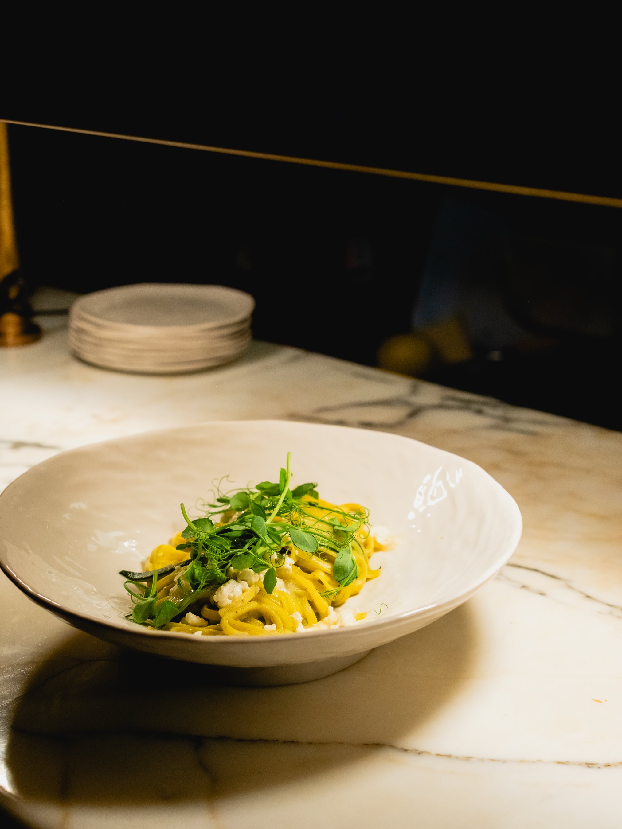 A white bowl with pasta topped with green herbs and microgreens, on a marble countertop.