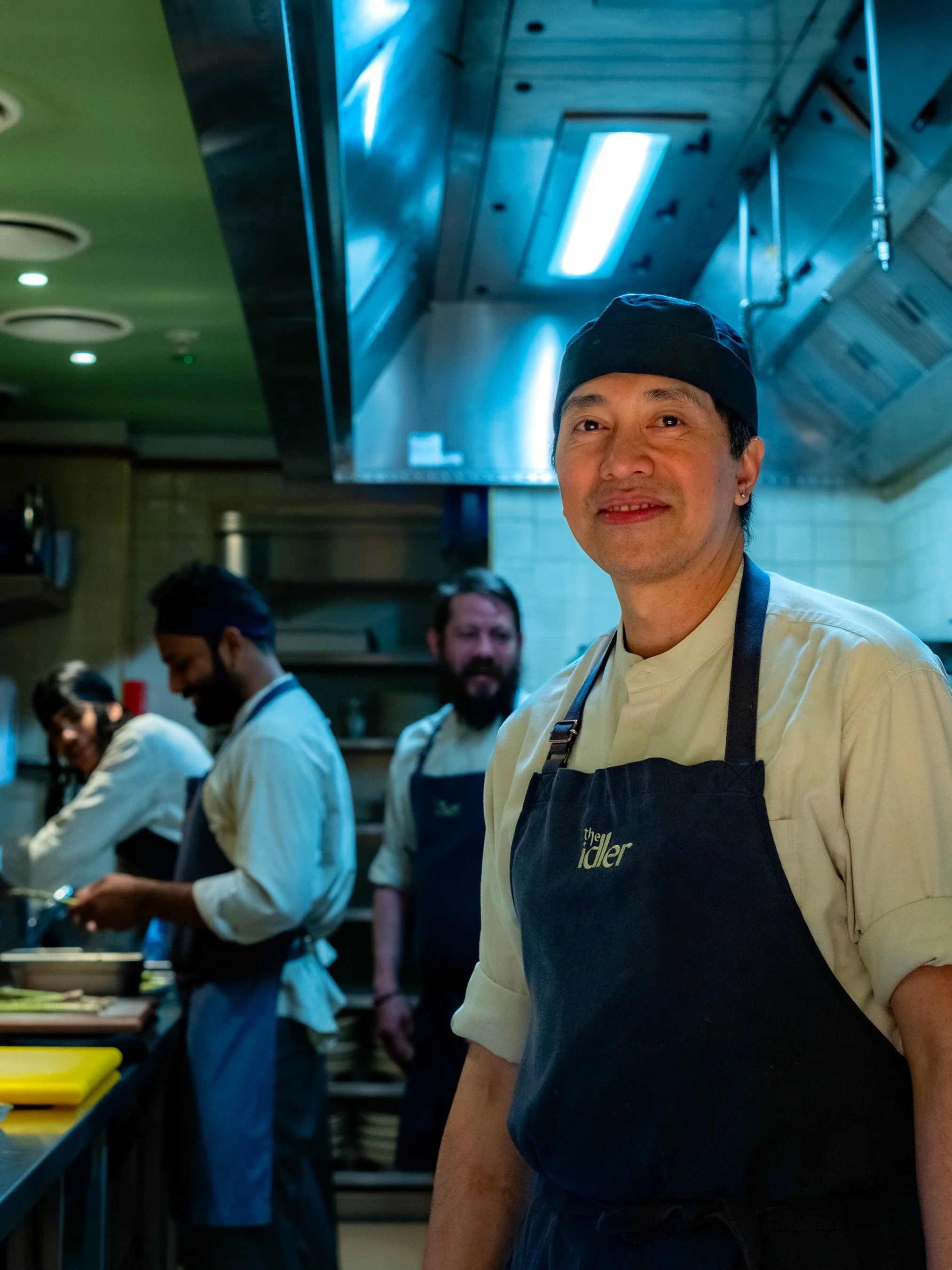 Chef wearing a black apron and hat in a busy kitchen, with other staff preparing food in the background.