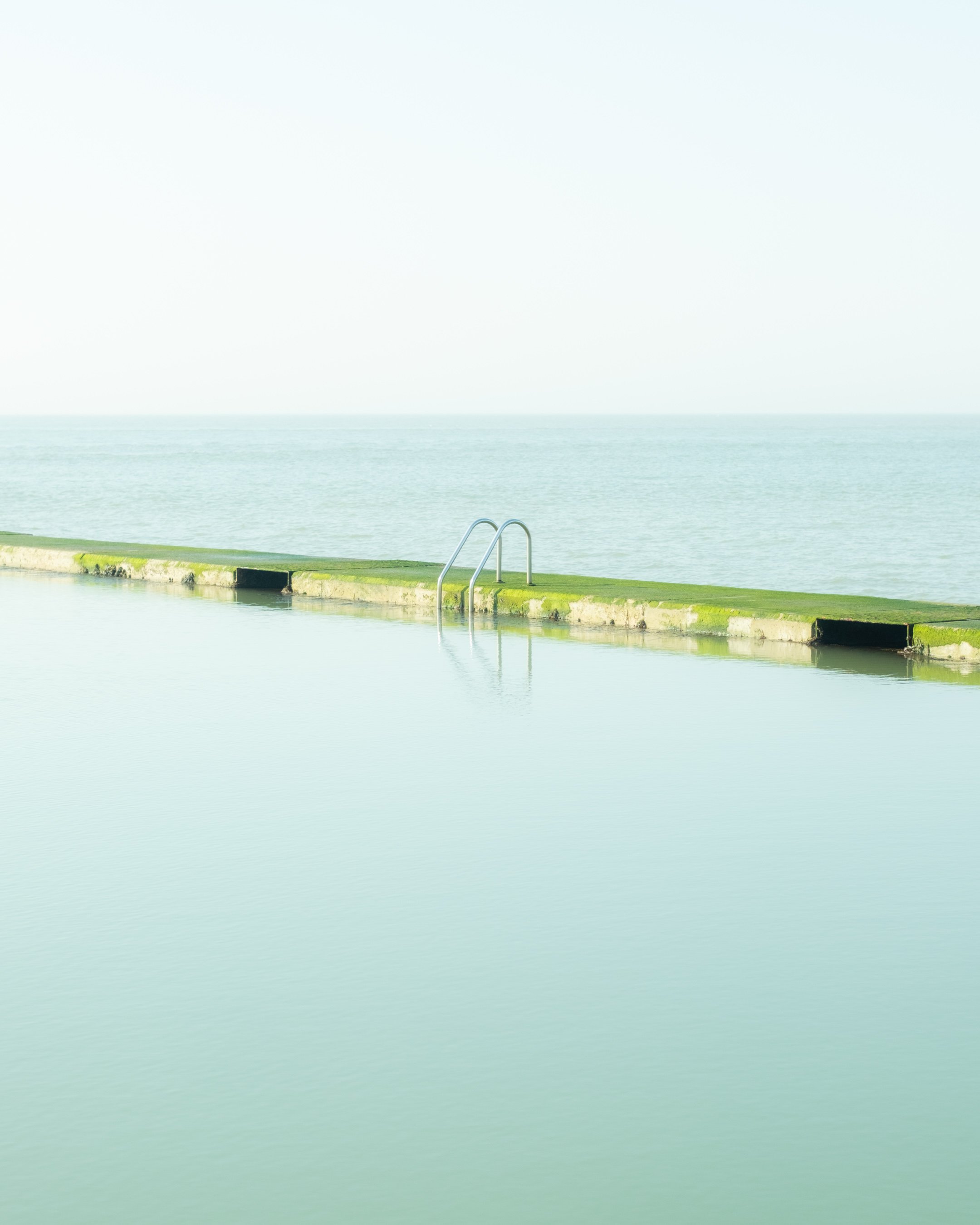 View of a calm body of water, possibly a lake or the ocean, with a concrete edge and metal steps leading into the water in the foreground. The sky is overcast or foggy, creating a tranquil and muted atmosphere.