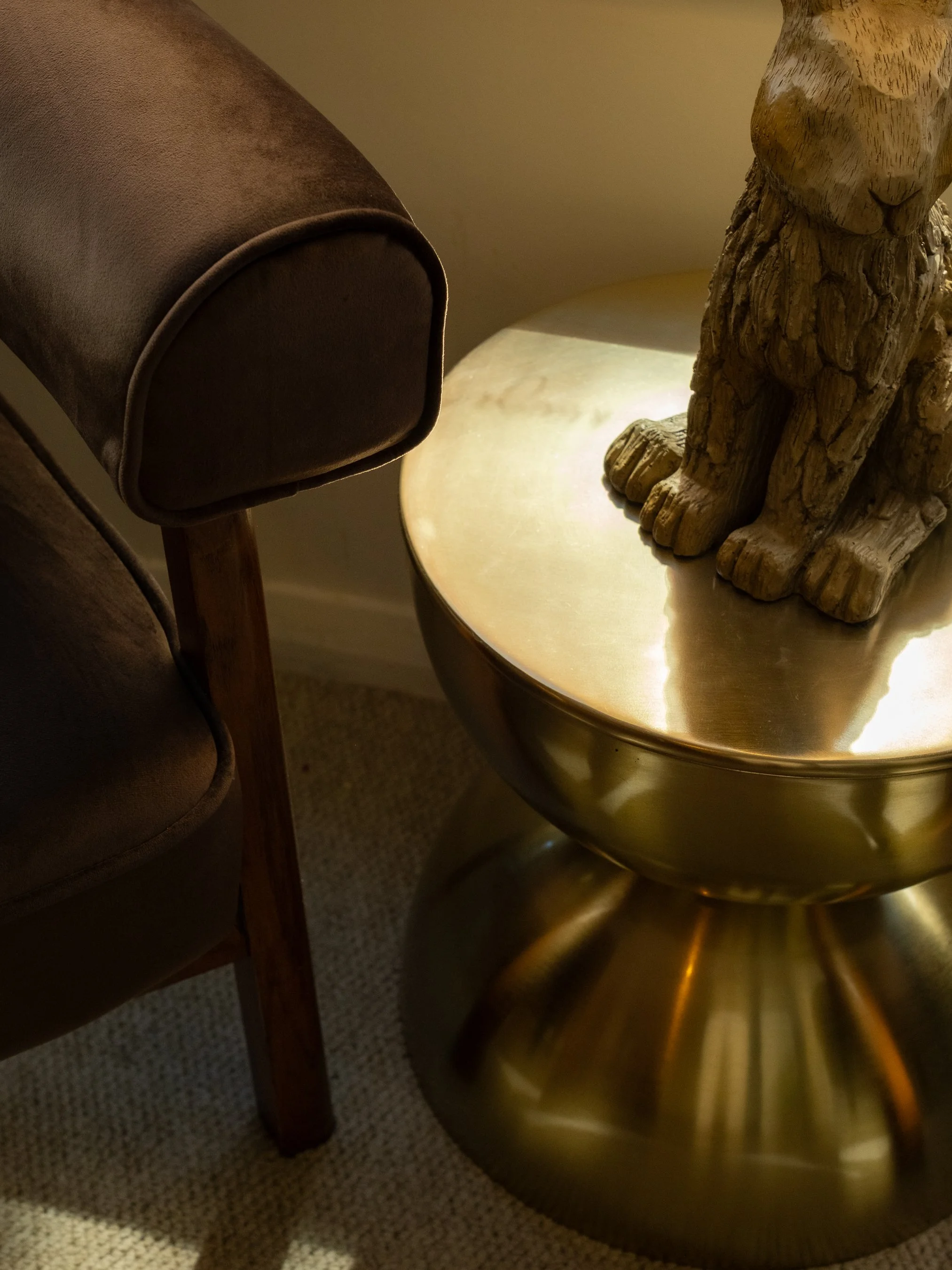 Close-up of a brown upholstered chair with wooden armrest next to a round, gold-colored metal side table featuring a wooden lion sculpture on top.