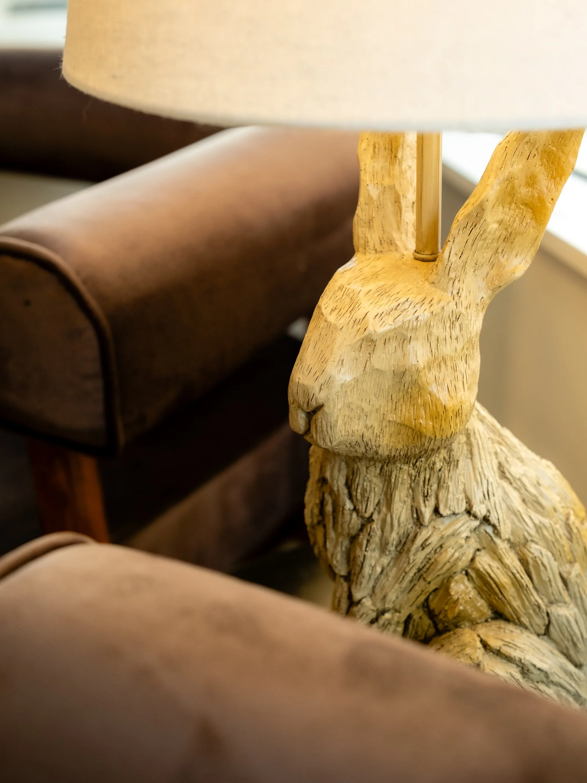 Close-up of a wooden rabbit sculpture with carved details, partially obscured by a beige fabric in the foreground, and a brown couch in the background.