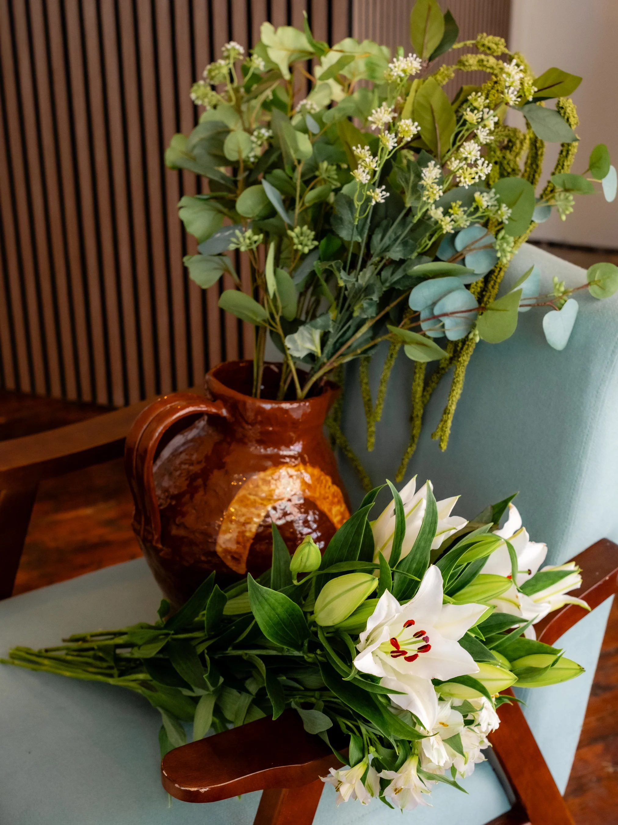 A bouquet of white lilies and green foliage resting on a wooden chair with a light blue cushion, next to a clay vase with green leaves and small white flowers. In the background, there is a wooden slat wall.