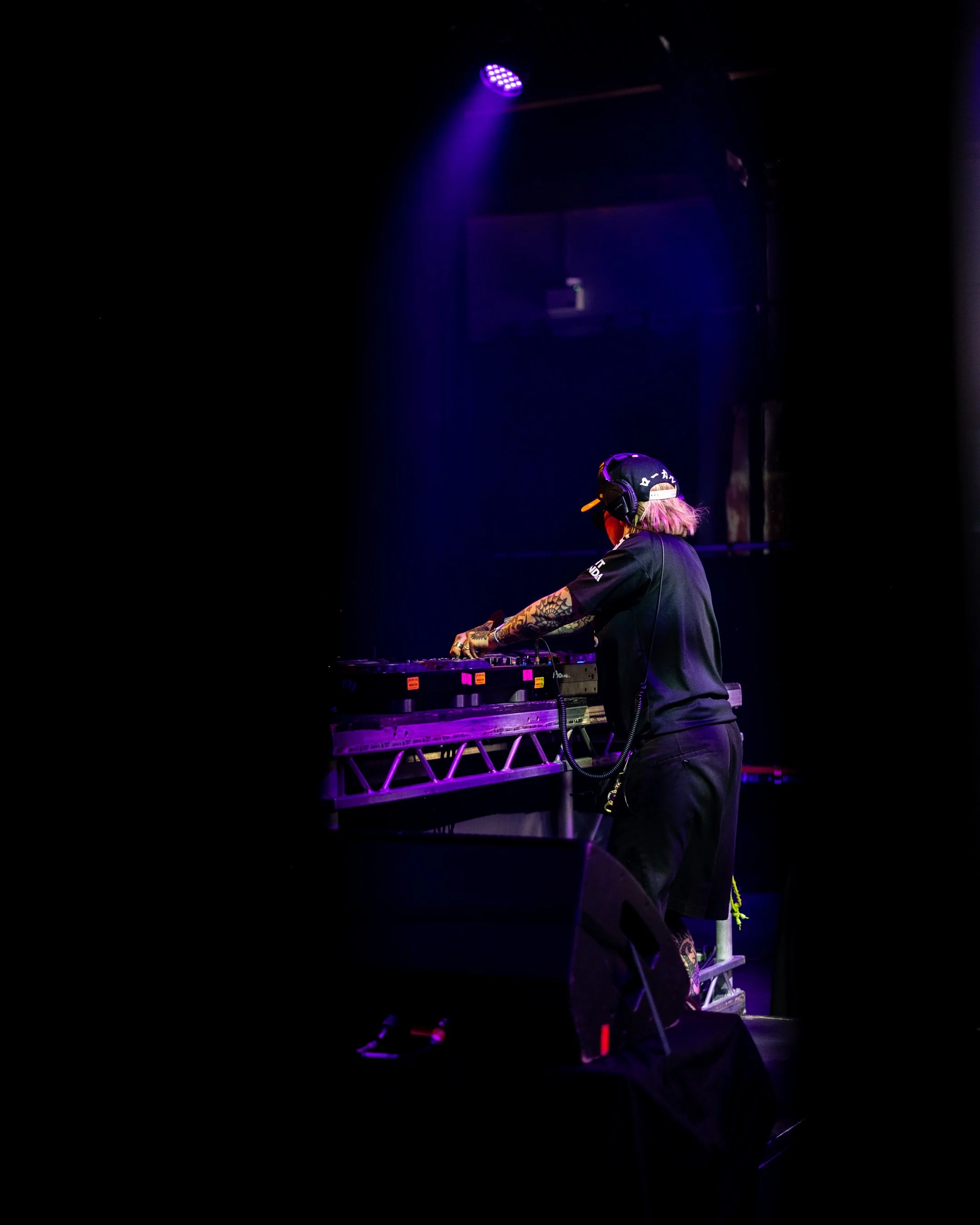A DJ with pink hair and tattoos on their arms is playing music on a DJ controller on stage, illuminated by purple and blue stage lights, with a dark background.