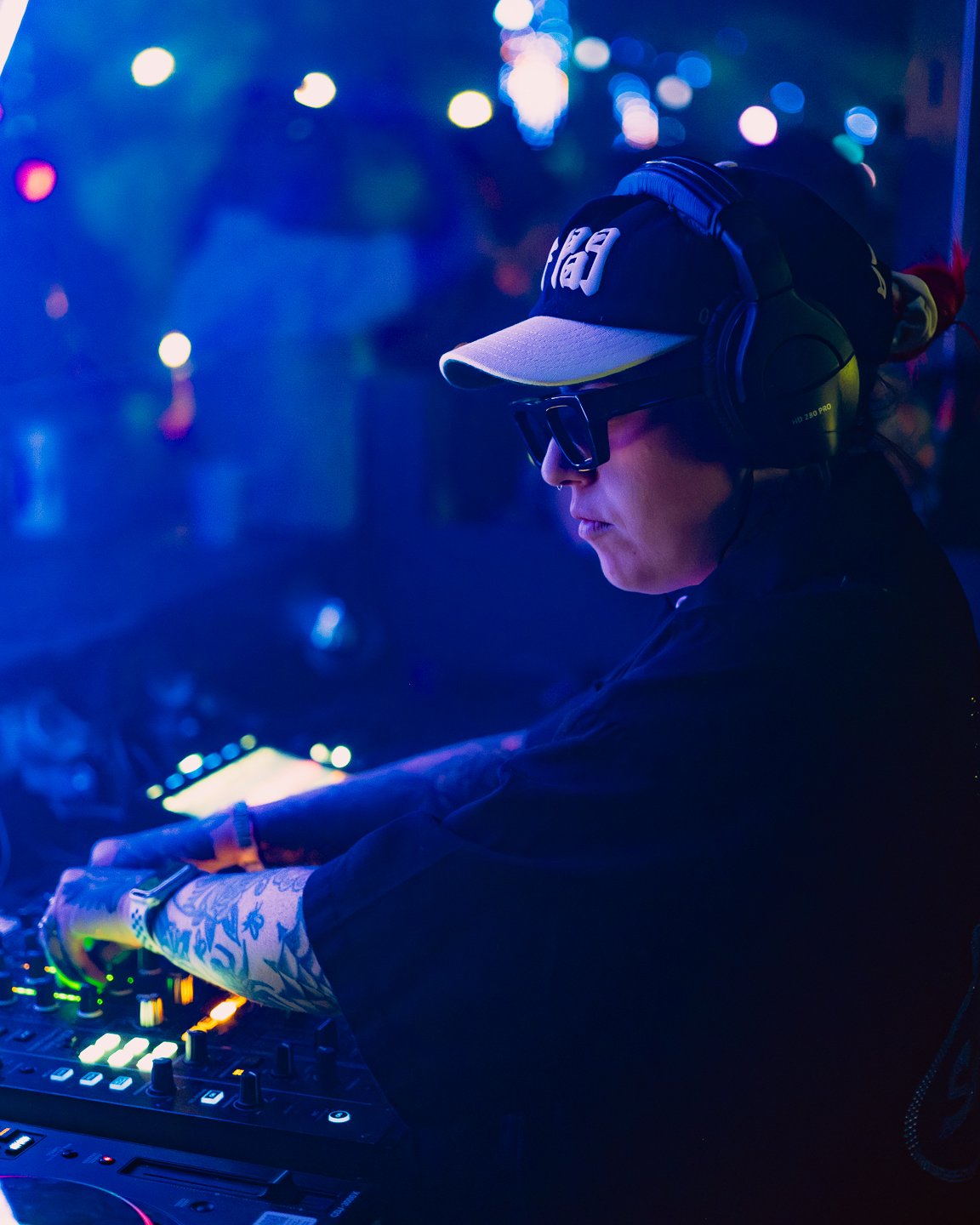 A person wearing sunglasses, a gray baseball cap, and headphones DJing at a nightclub with colorful lights in the background.