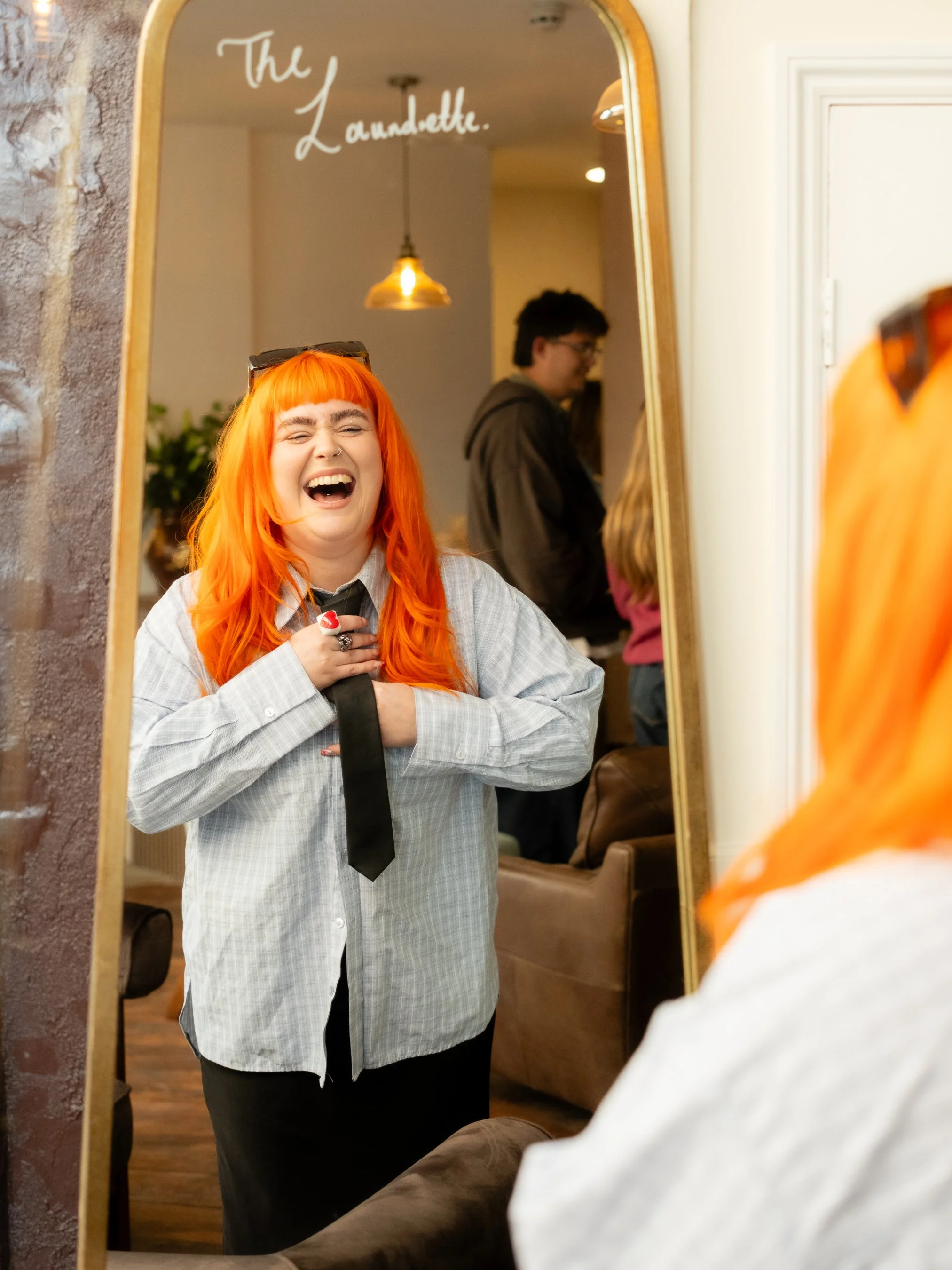 A woman with bright orange hair, wearing a gray shirt, black tie, and glasses on her head, laughing and holding her chest. She is looking into a mirror that has the words 'The Laundrette' written on it. A man in a dark hoodie is in the background, us