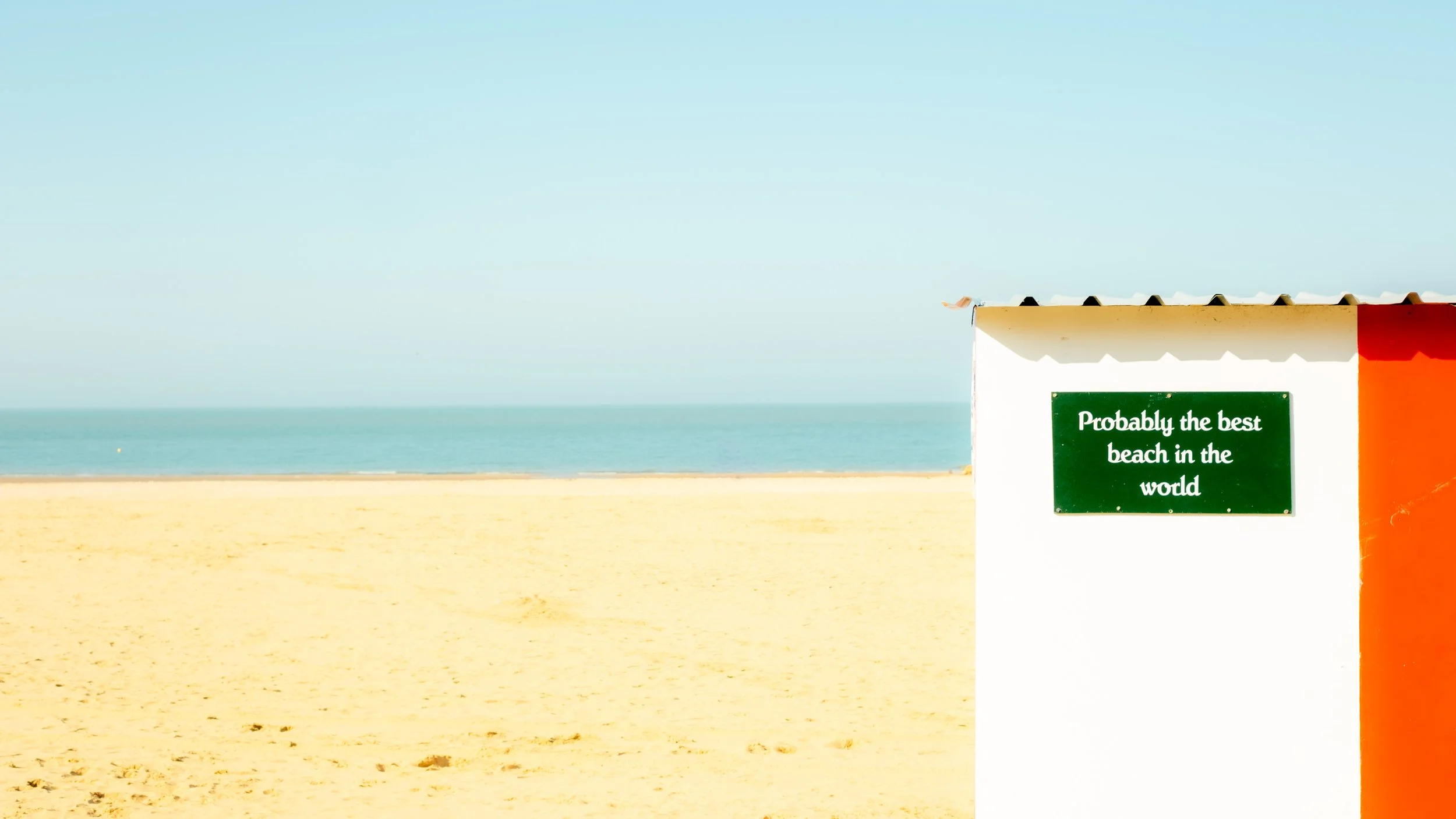 Beach scene with sandy shore, calm ocean, and clear sky. A beach hut with a sign that reads 'Probably the best beach in the world.'