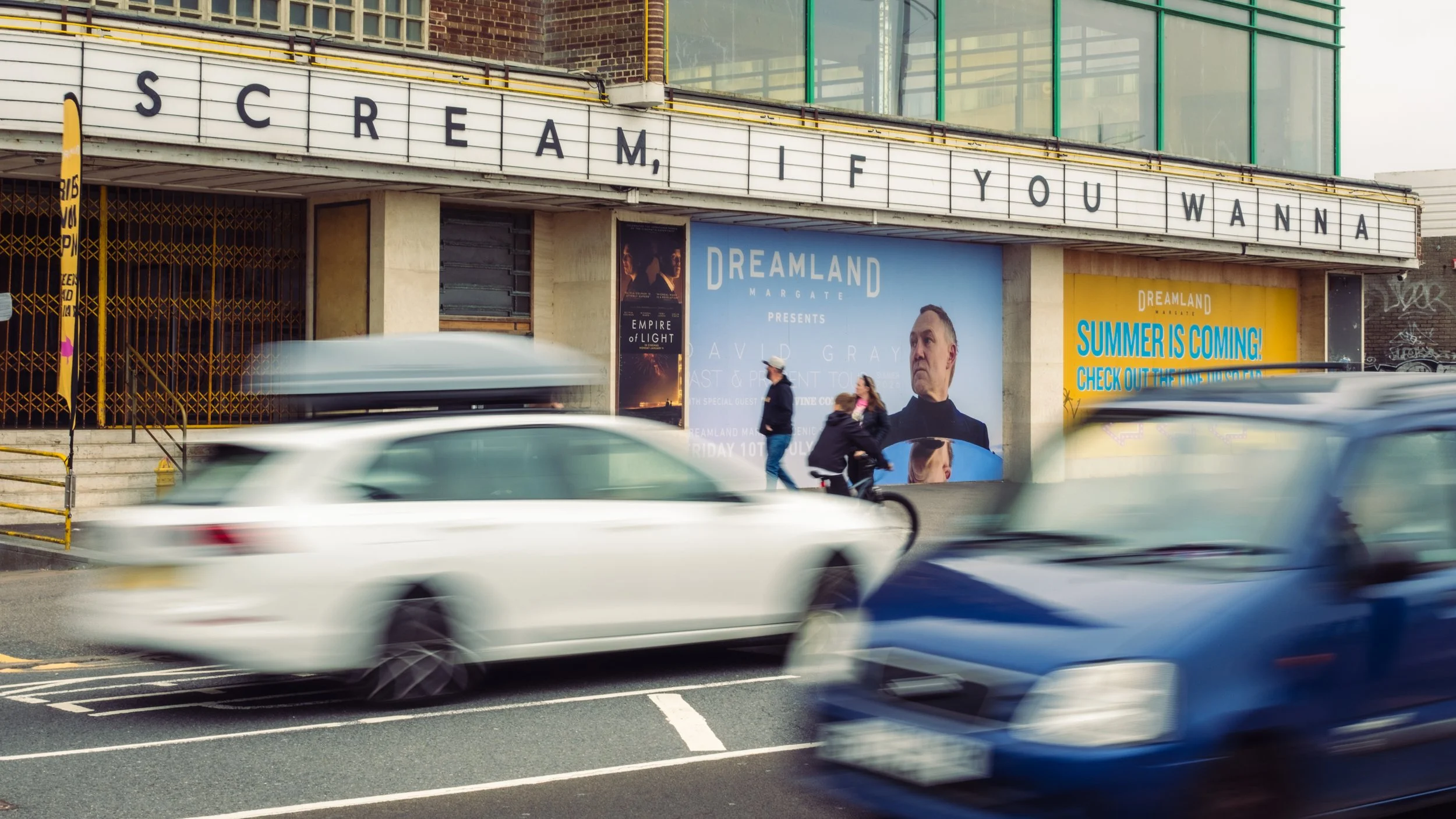 Street scene with moving cars, pedestrians, and a theater marquee displaying the message "I SCREAM, IF YOU WANN A" and advertising posters for the show "Dreamland" featuring David Gray.