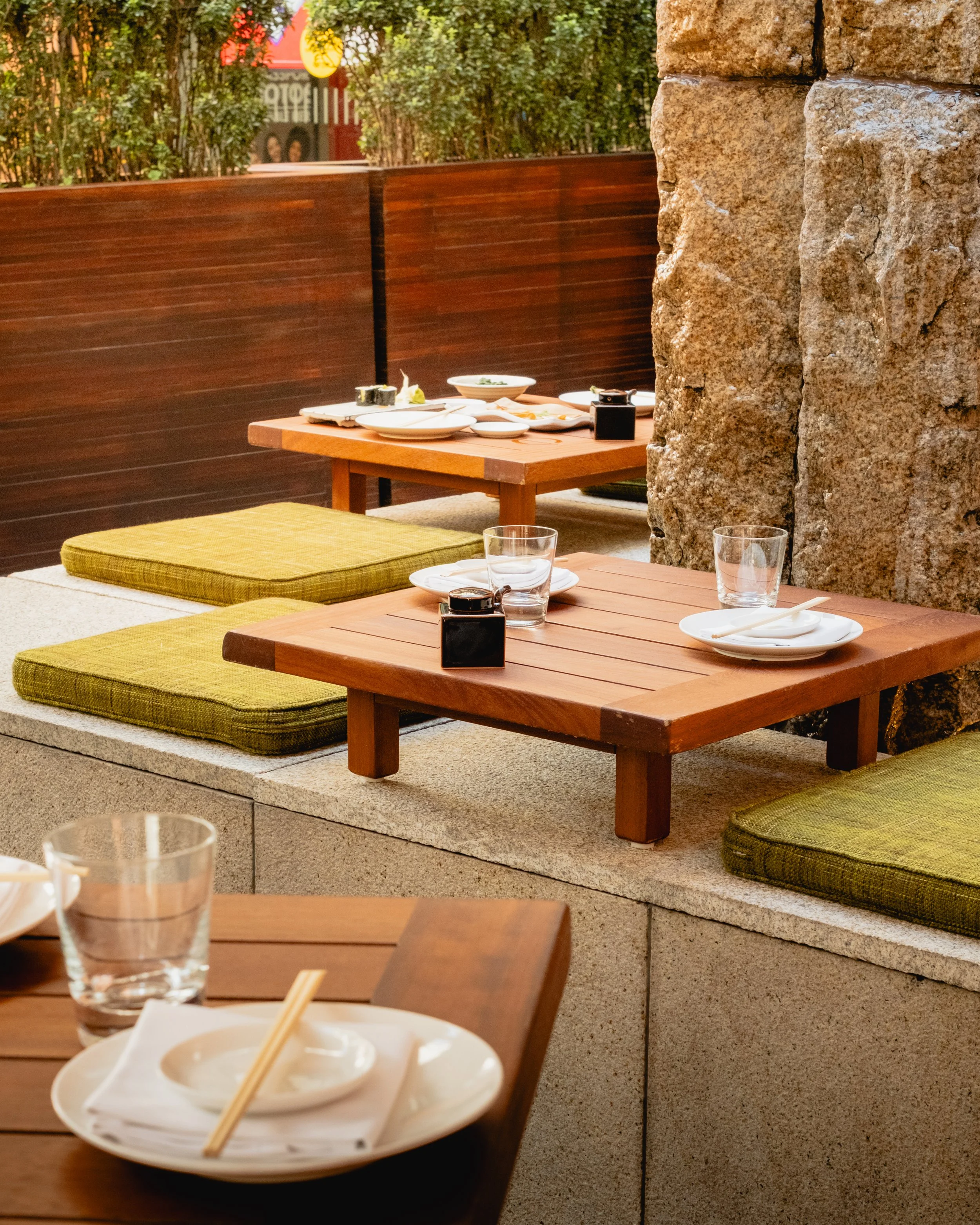 An outdoor dining area with wooden tables, green cushions, and stone walls. Plates, chopsticks, and glasses with water are on the tables, with a background of bushes and a sign with bright colors.