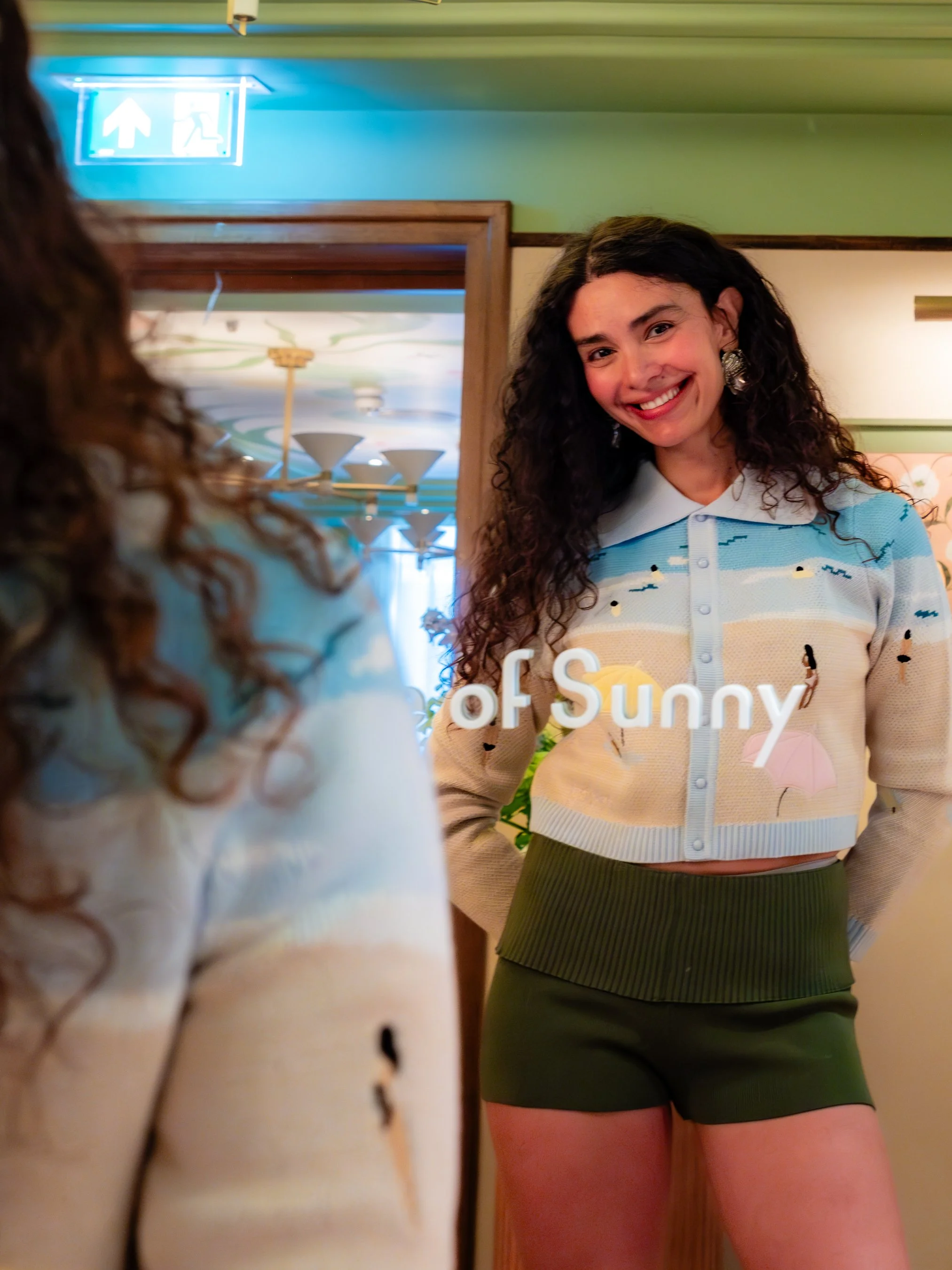 A woman with long curly hair is smiling and standing in front of a mirror, wearing a colorful striped sweater and green shorts. An emergency exit sign is visible at the top of the image.