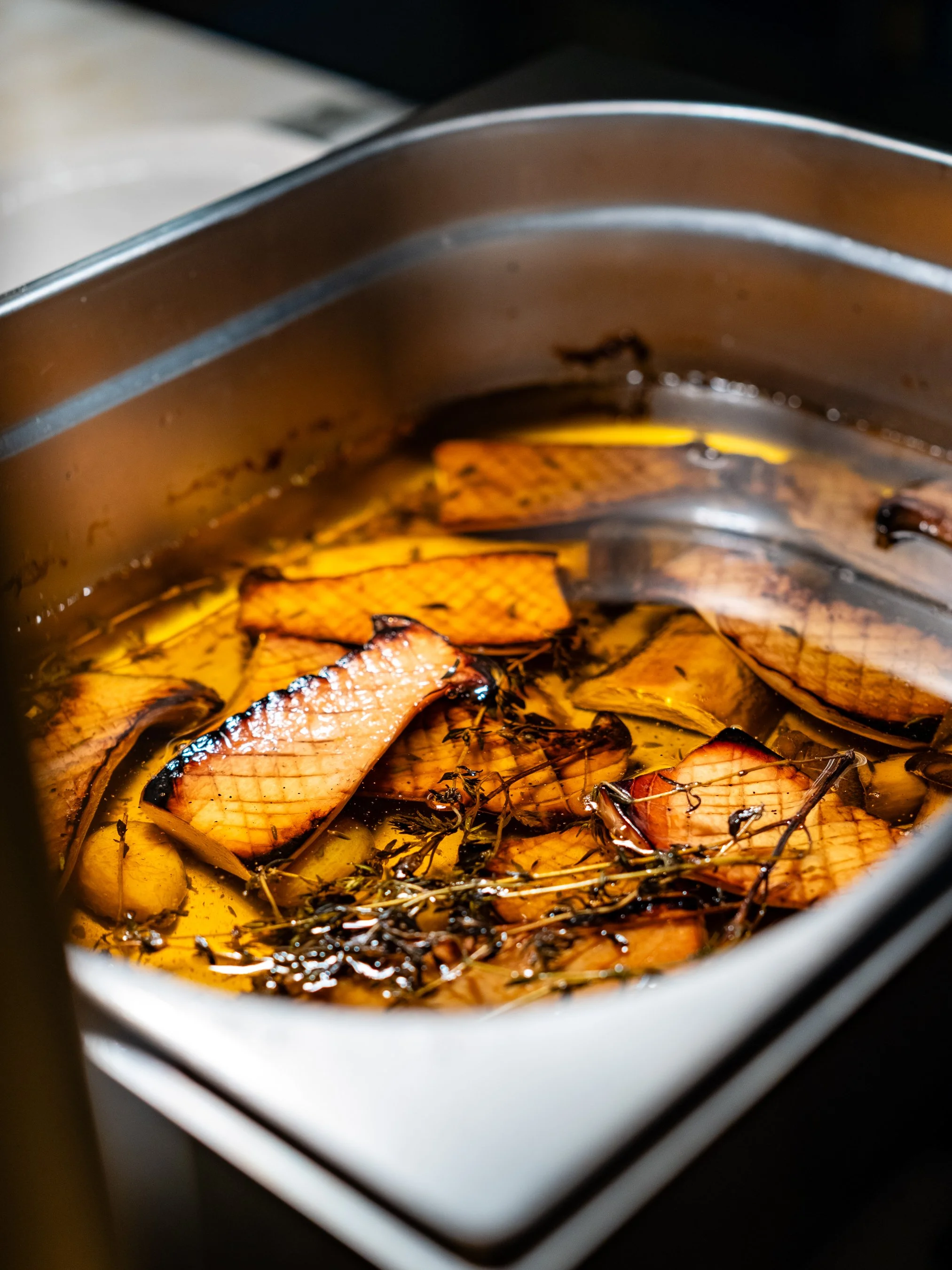 Close-up of a cooking pot containing burnt fish fillets and herbs simmering in a dark, oily broth.