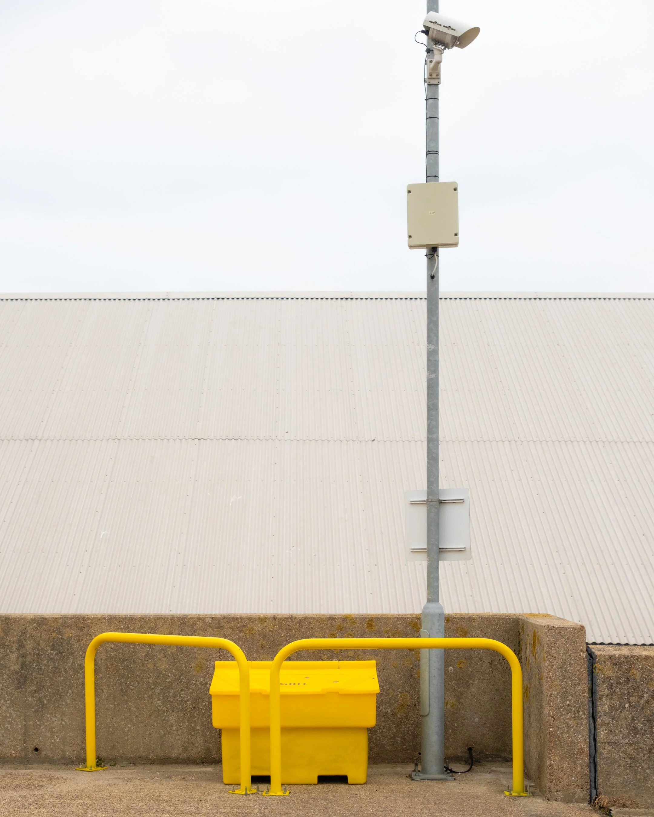 A yellow recycling bin with a yellow barrier in front of an industrial building wall. There is a grey pole with a camera and a beige box attached above the bin. The building wall is white with a corrugated pattern.