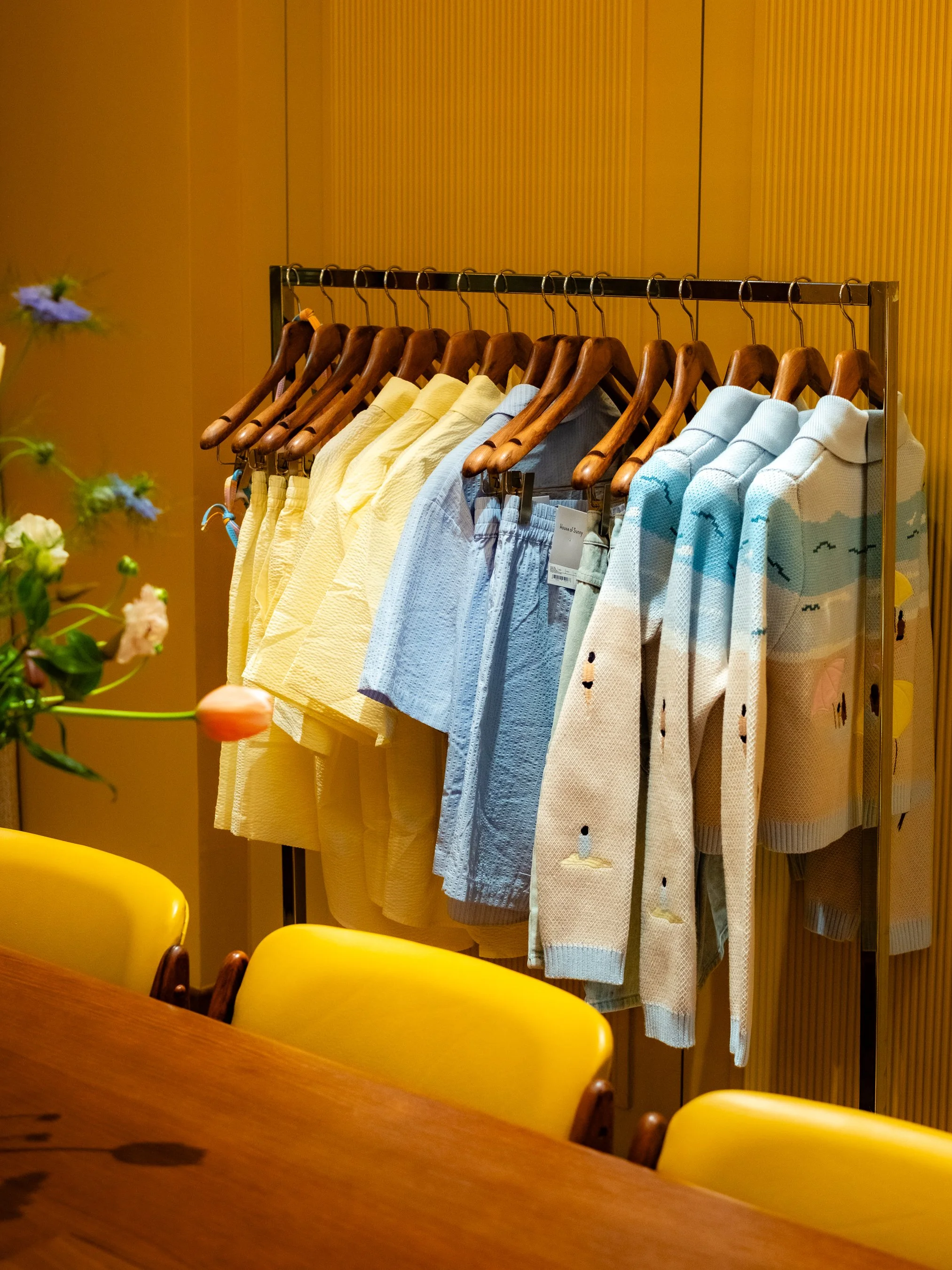 Clothing rack with yellow, blue, and white shirts and sweaters, background of wood-paneled wall.
