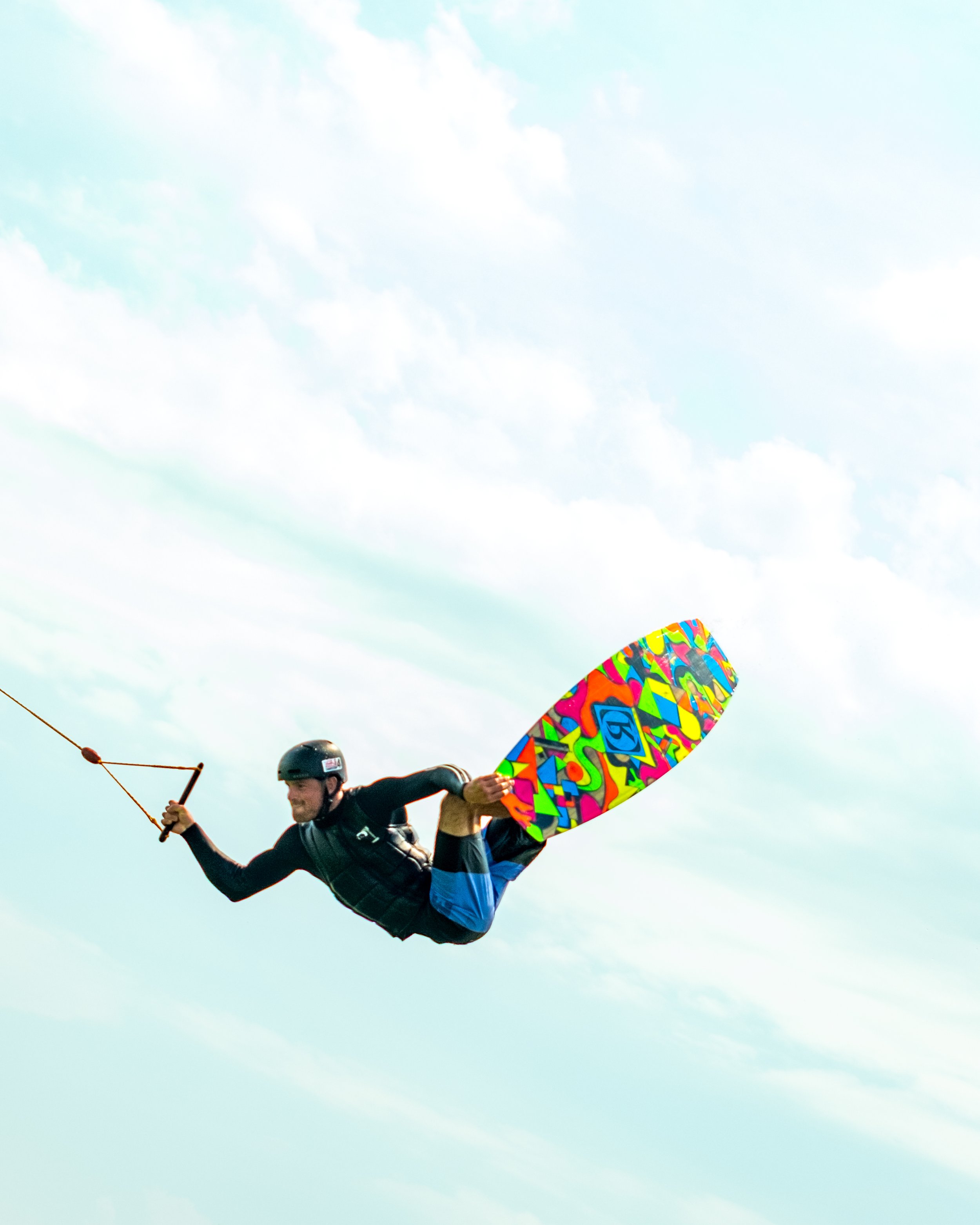 A person in mid-air wakeboarding with a colorful, abstract-designed wakeboard, holding onto a rope, against a blue sky background with clouds.